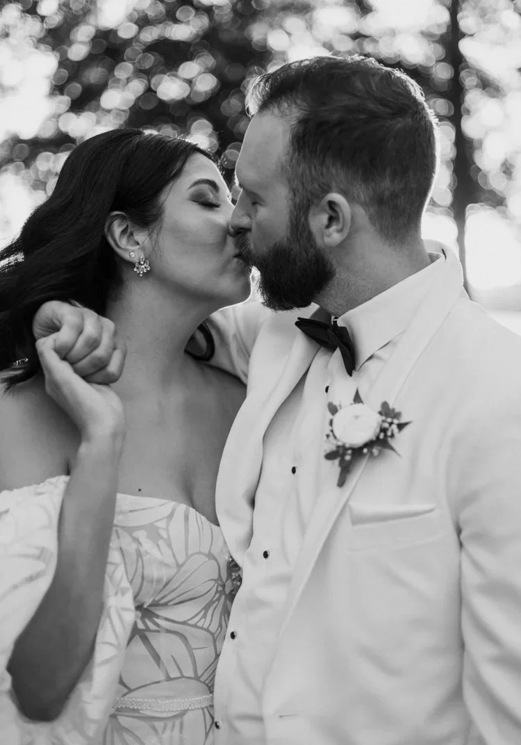 A black and white photo of a couple kissing outdoors, dressed in formal wedding attire, with the woman wearing earrings and an off-shoulder dress, and the man wearing a tuxedo with a boutonniere.