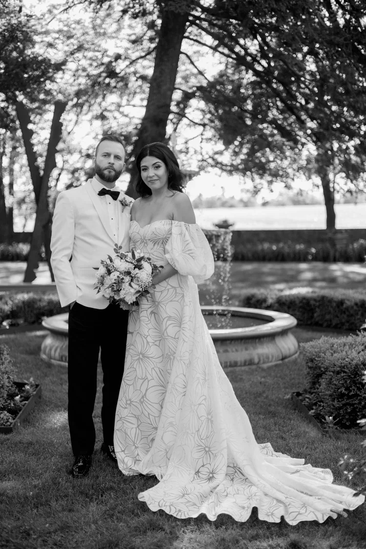A black-and-white photo of a bride and groom standing outdoors by a fountain, surrounded by trees and landscaping. The bride wears an off-shoulder wedding gown with a floral pattern and holds a bouquet, while the groom is in a white tuxedo with a bla