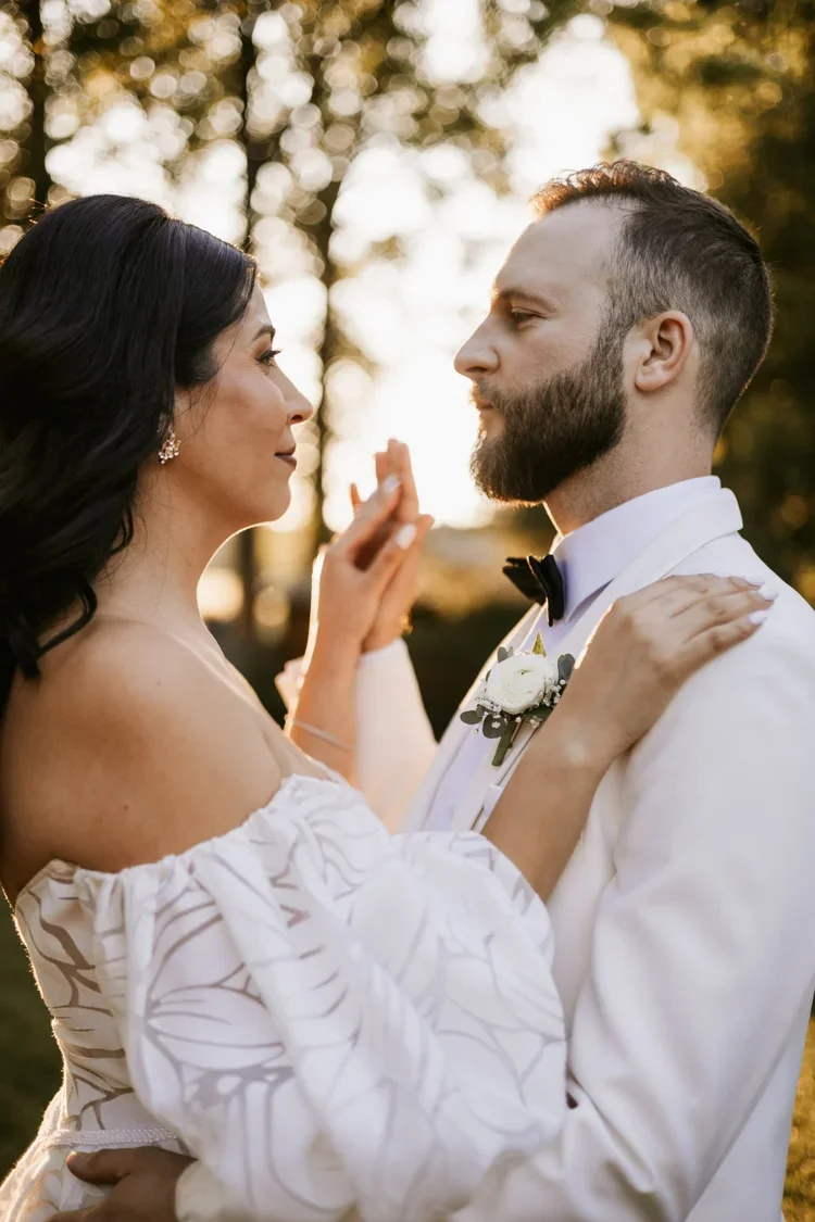 A bride and groom facing each other outdoors during sunset, with the bride's hands gently touching the groom's face, both in wedding attire, surrounded by trees.