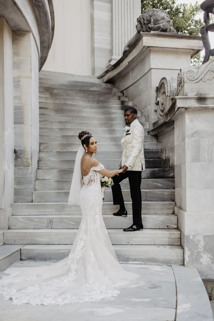 A bride and groom holding hands on stone steps outside a historic building with classical architecture and statues, during their wedding.