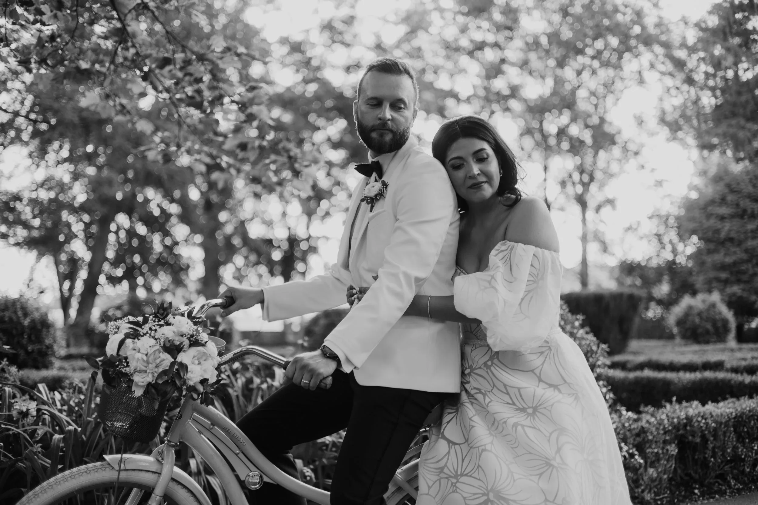 A black-and-white photo of a newlywed couple posing outdoors. The groom is sitting on a bicycle decorated with flowers, and the bride is leaning on his back, resting her head on his shoulder.