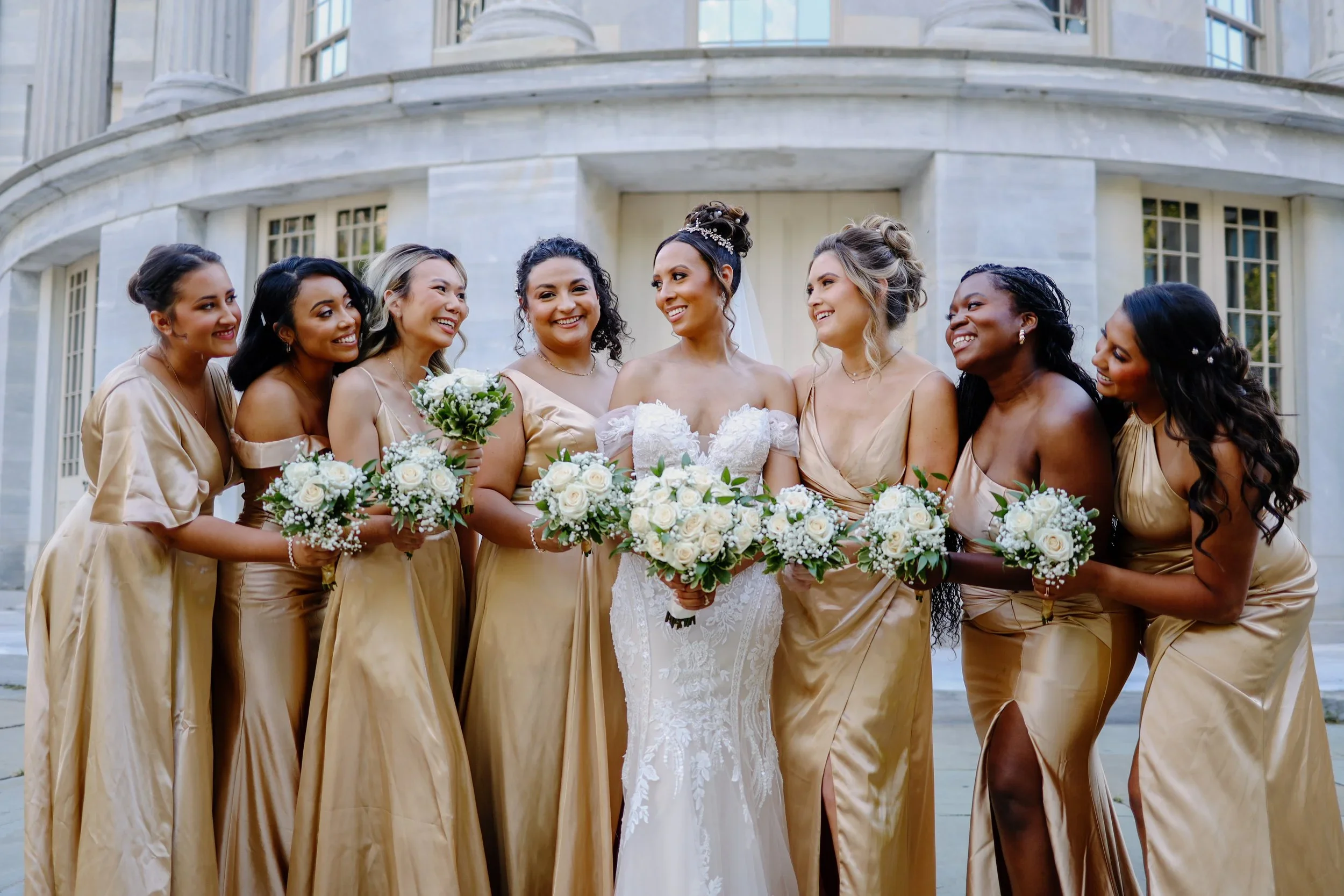 Bride in white wedding dress surrounded by eight bridesmaids in matching gold dresses, holding bouquets of white roses, standing outside a large stone building, smiling and looking at each other.