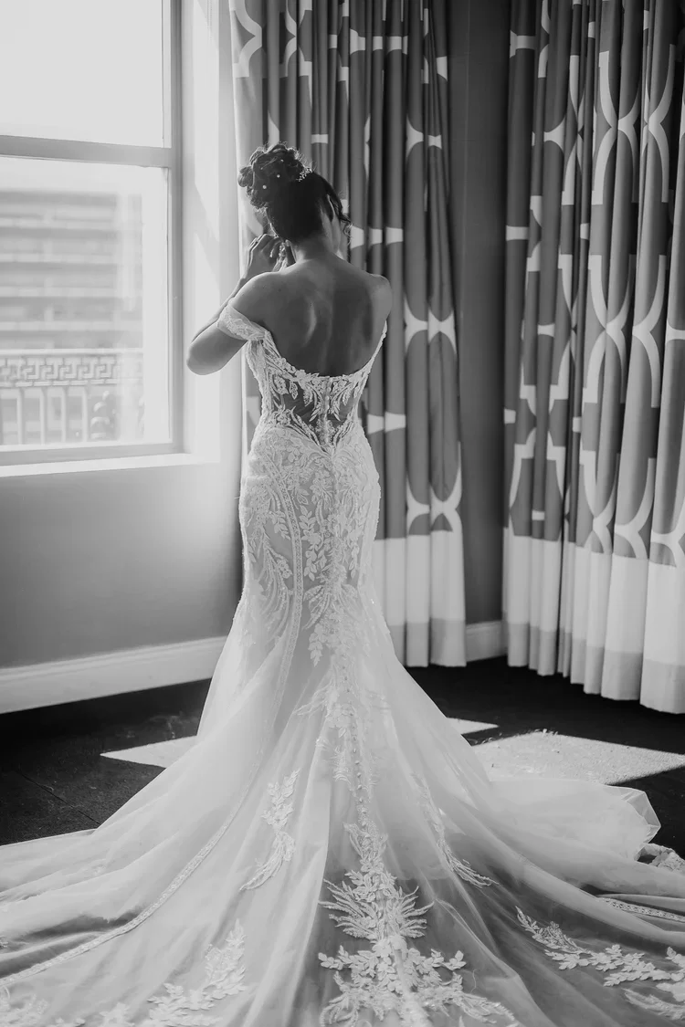 A woman in a lace wedding gown standing indoors near a window with curtains, back to the camera, adjusting her hair.