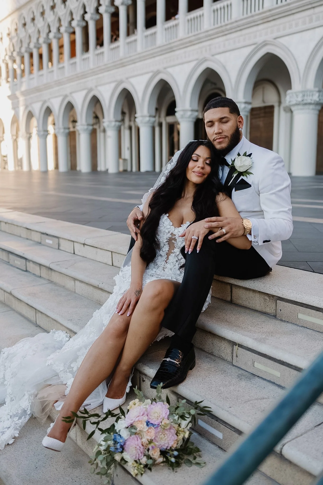 Bride and groom in wedding attire sitting on steps outside a building with arches, the bride leaning on the groom, both with peaceful expressions. A bouquet of pink, purple, and blue flowers is placed on the steps beside them.