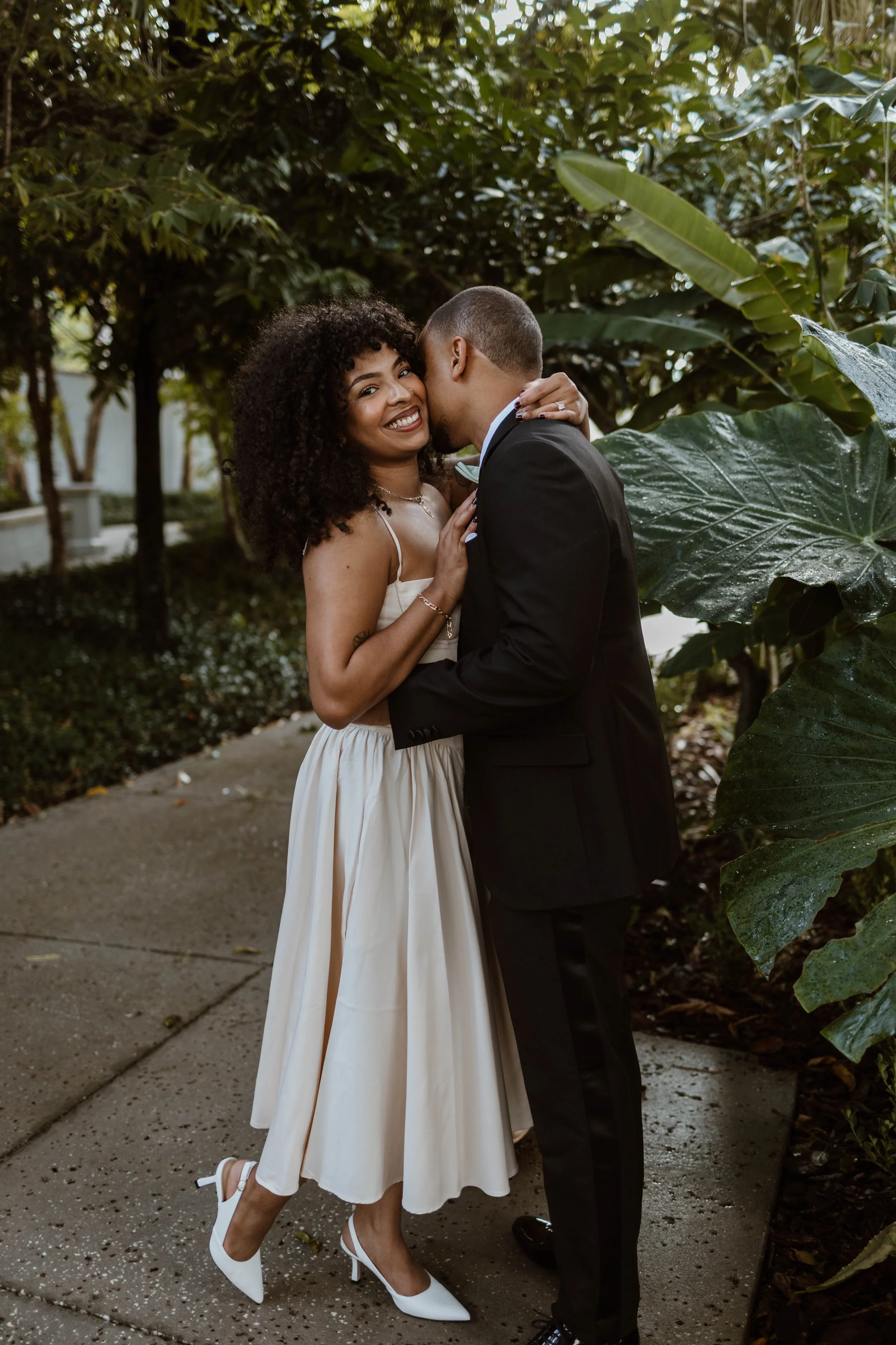 A couple dressed in formal attire embracing outdoors amidst lush green foliage.