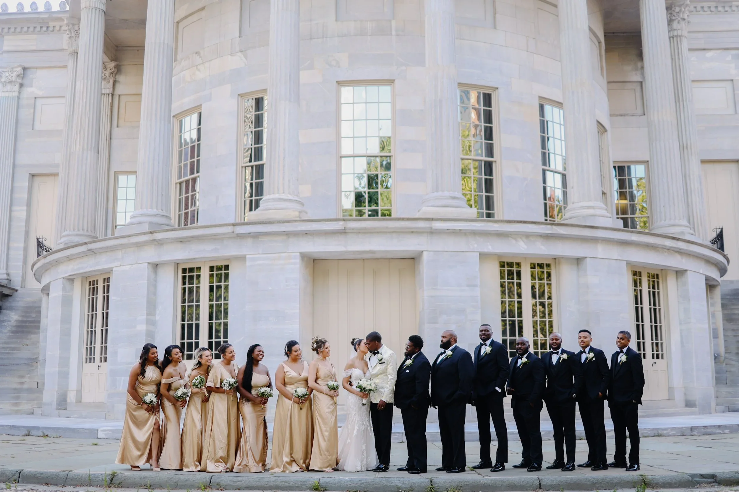 A wedding party outside a classical building with large columns. The bride and groom are kissing in the center, surrounded by bridesmaids in gold dresses on one side and groomsmen in black suits on the other.