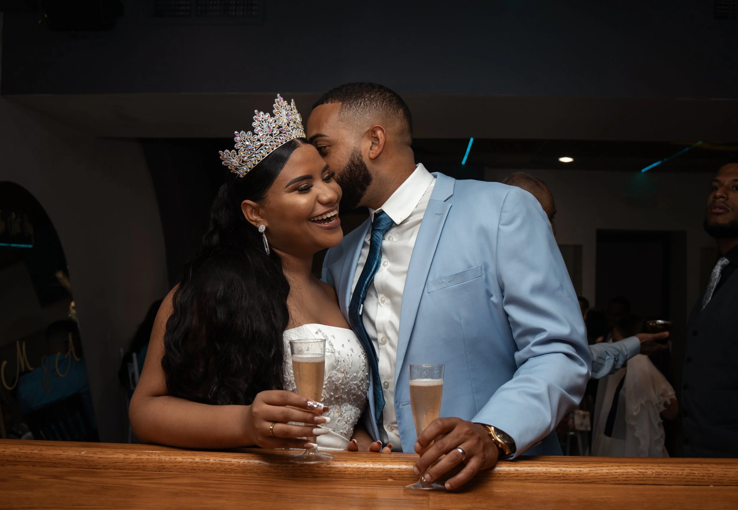 A bride and groom celebrating at their wedding, with the bride wearing a tiara and holding a glass of champagne, and the groom whispering in her ear while also holding a glass of champagne. They are smiling and standing behind a wooden bar.