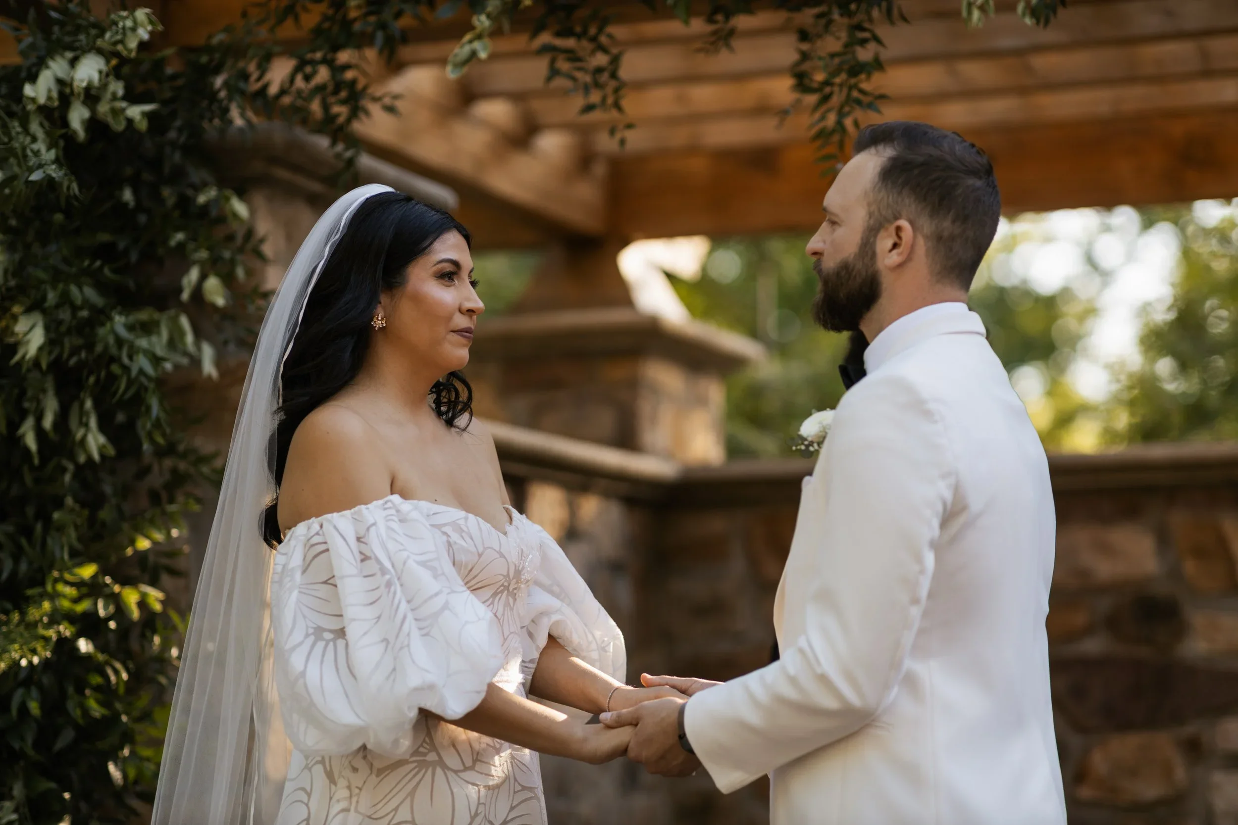 A bride and groom holding hands during their wedding ceremony outdoors, with the bride wearing an off-the-shoulder white dress and veil, and the groom in a white tuxedo jacket, standing in front of a wooden and stone backdrop.