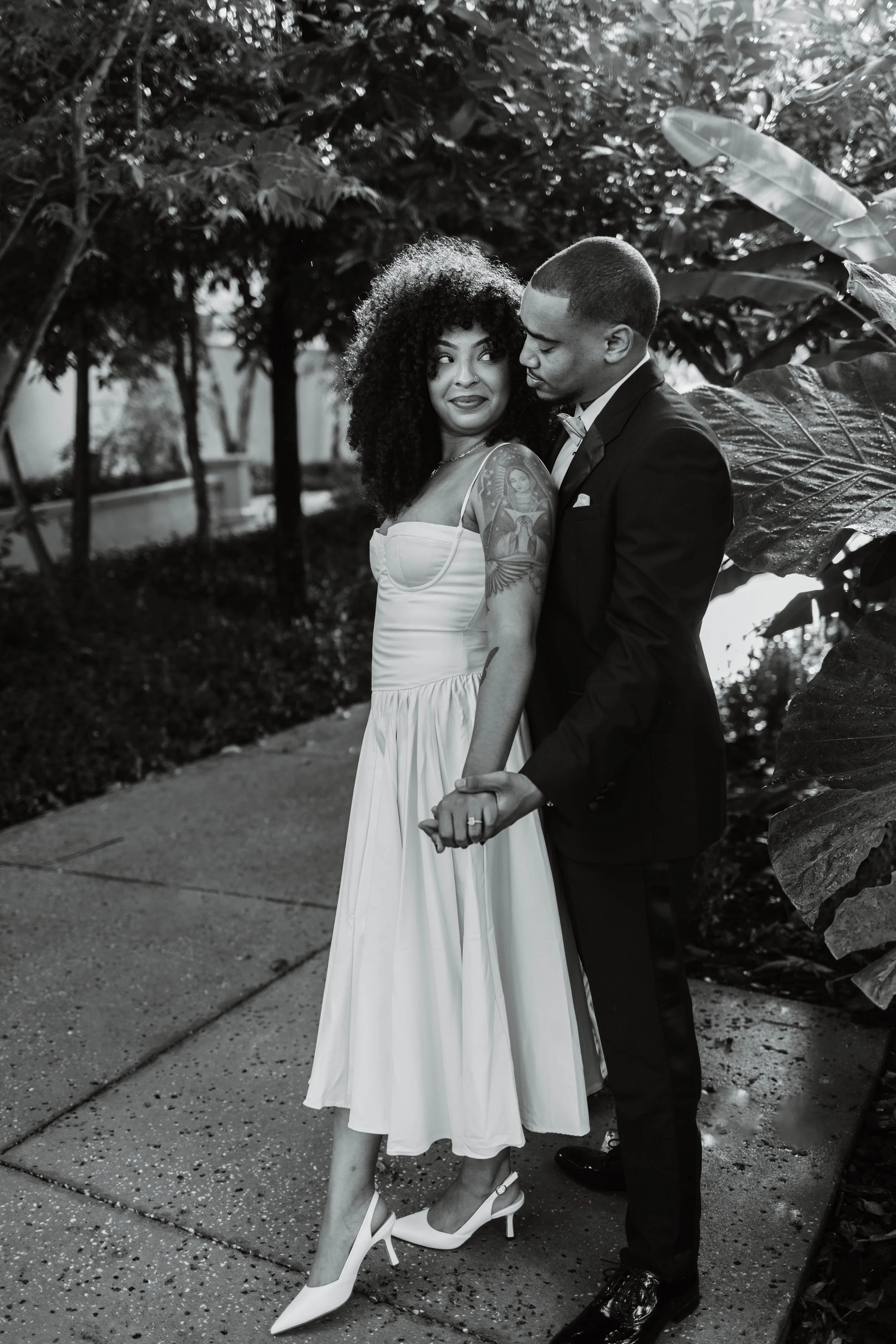 A Black and White photo of a couple holding hands, standing outdoors among trees, dressed formally with the woman in a dress and heels, and the man in a suit.
