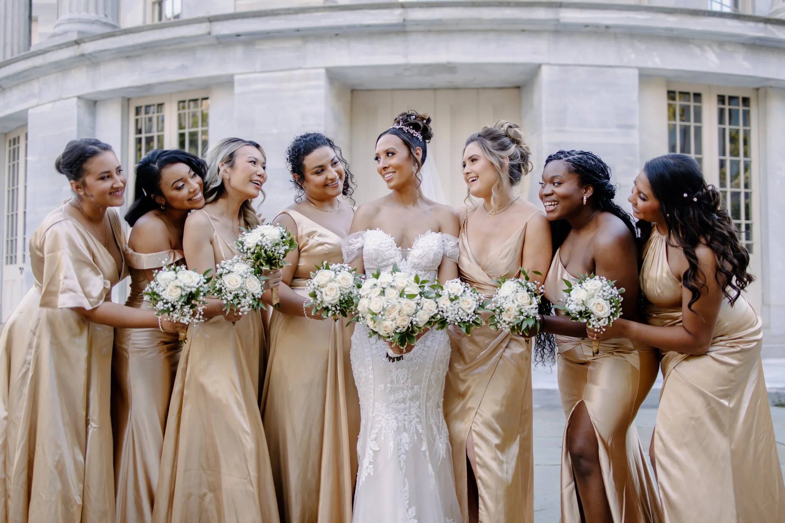 A bride and her bridesmaids standing outside in front of a white building. The bride is in a white lace wedding gown holding a bouquet, surrounded by bridesmaids in matching beige dresses holding bouquets, all smiling and looking at the bride.