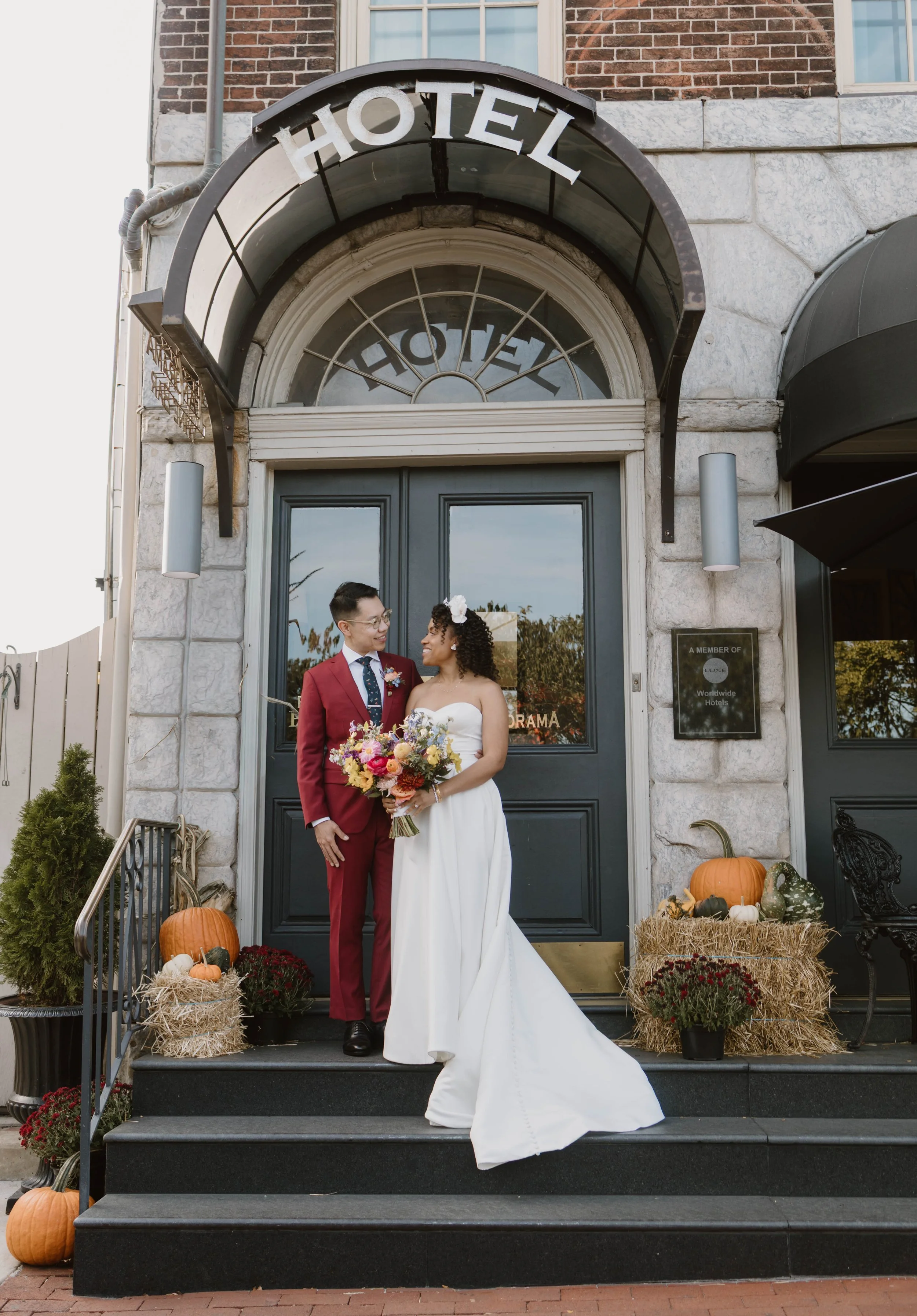 A newlywed couple stands on the steps of a hotel decorated with fall pumpkins and hay bales. The groom, wearing a red suit, white shirt, and glasses, looks at the bride, who is dressed in a strapless white wedding gown with a large floral headpiece a