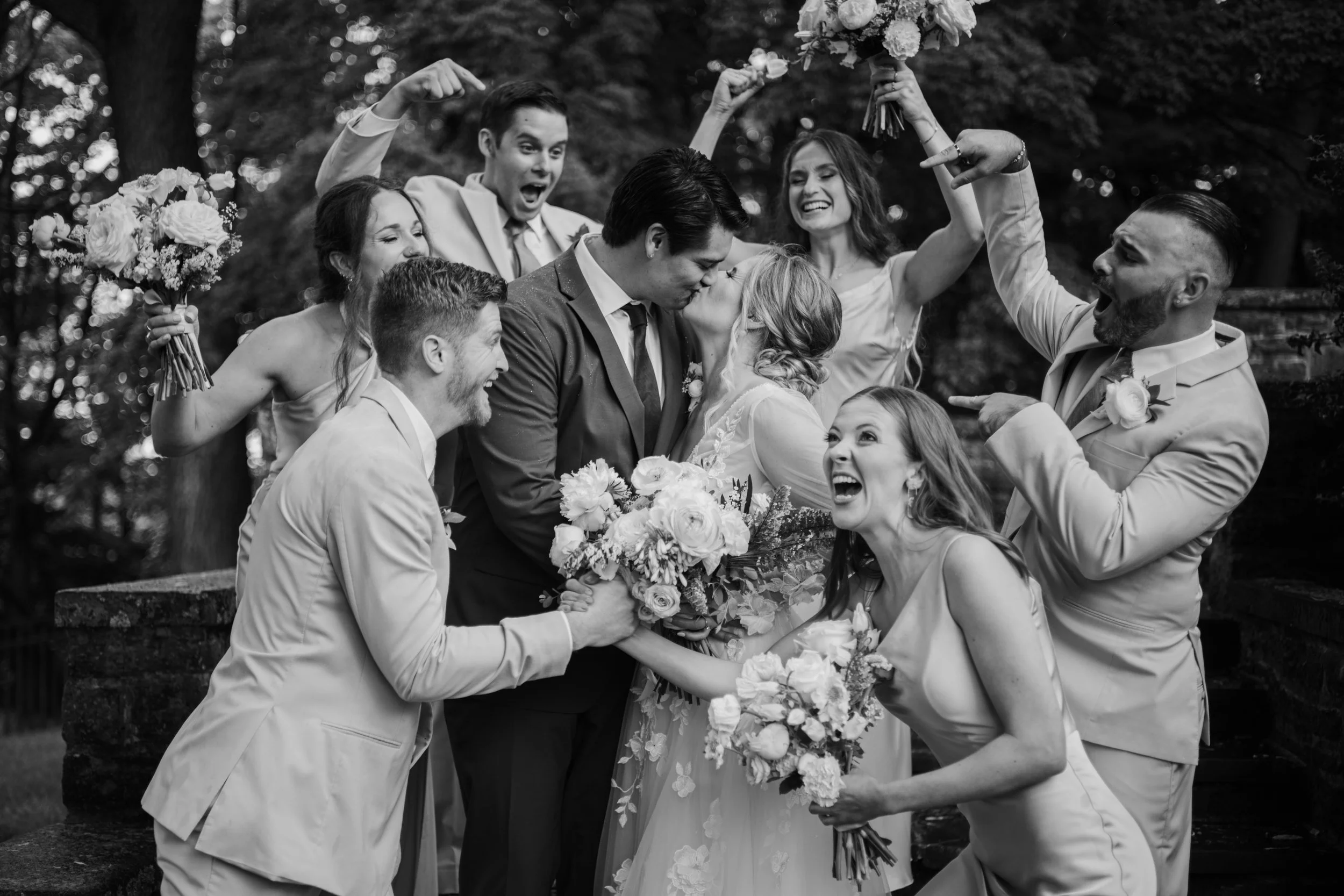 A black and white photo of a wedding celebration showing a bride and groom kissing surrounded by their wedding party, who are joyfully celebrating and holding bouquets, outdoors in a lush green area.