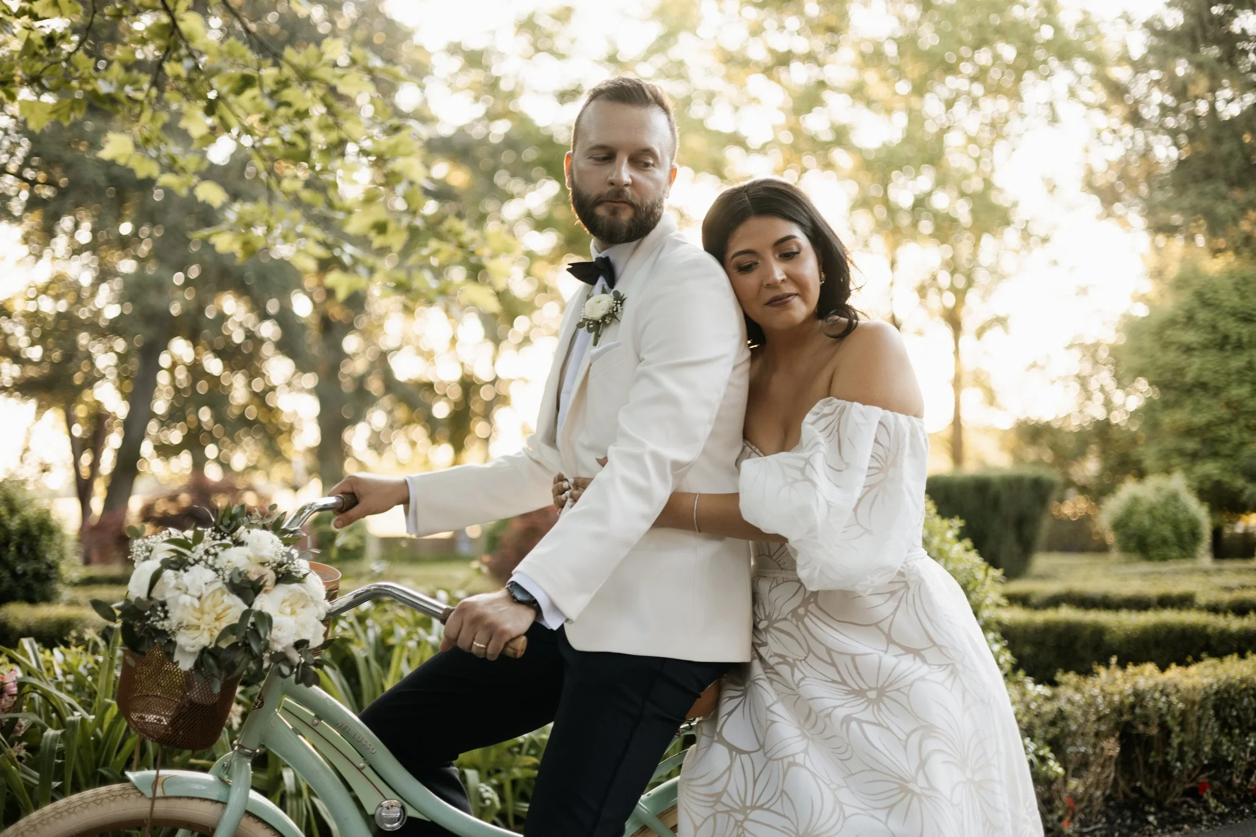 A bride and groom in wedding attire standing on a vintage bicycle decorated with flowers, outdoors in a park with trees and greenery in the background during sunset.