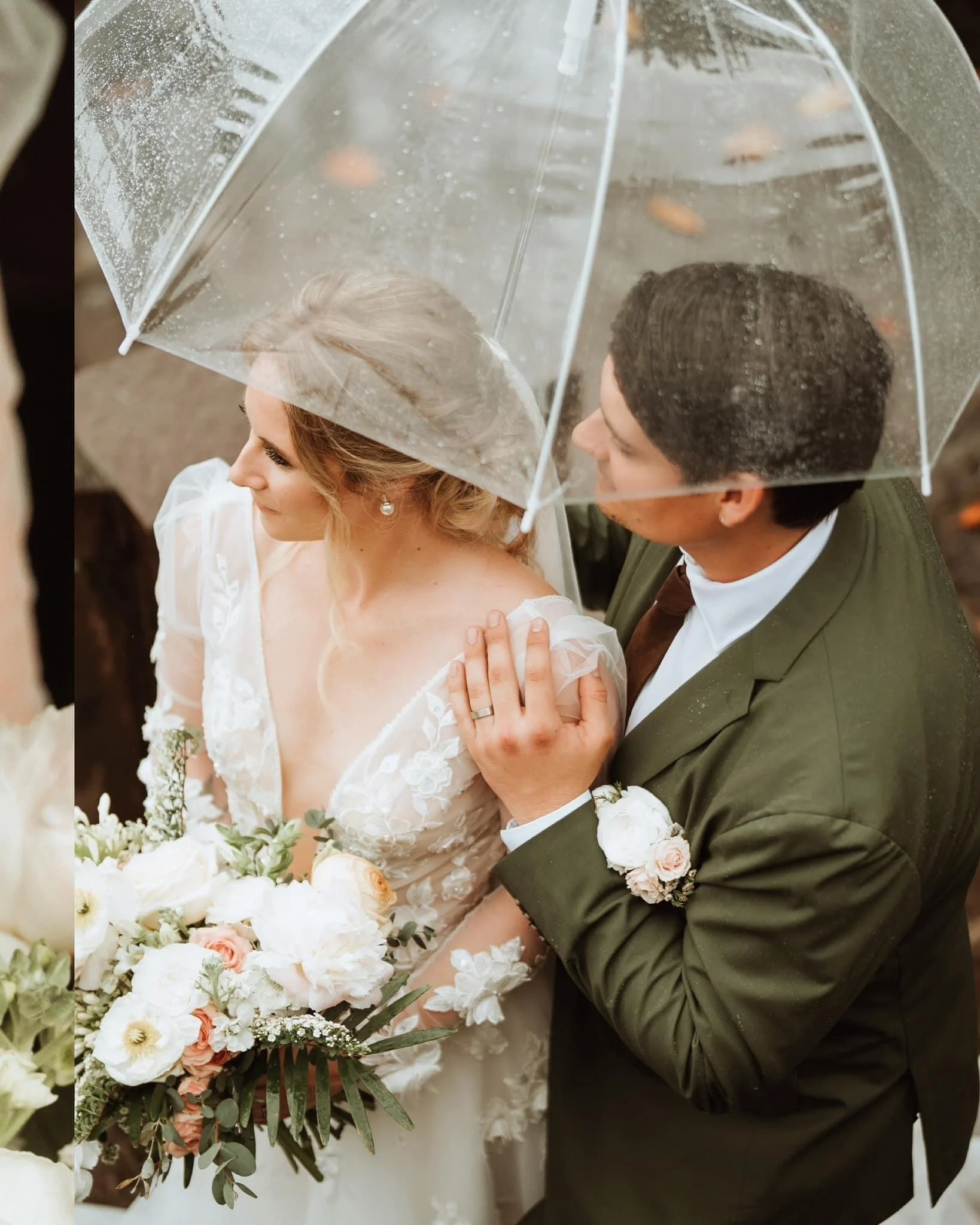 Bride and groom under a clear umbrella on their wedding day, with the bride holding a bouquet of white and blush flowers, rain visible on the umbrella.