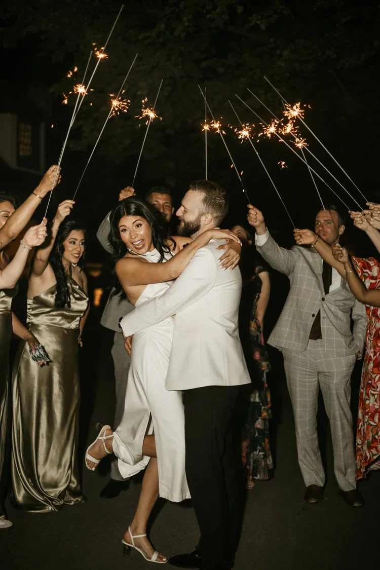 Couple dancing at a celebration with sparklers, surrounded by friends dressed in formal and party attire.
