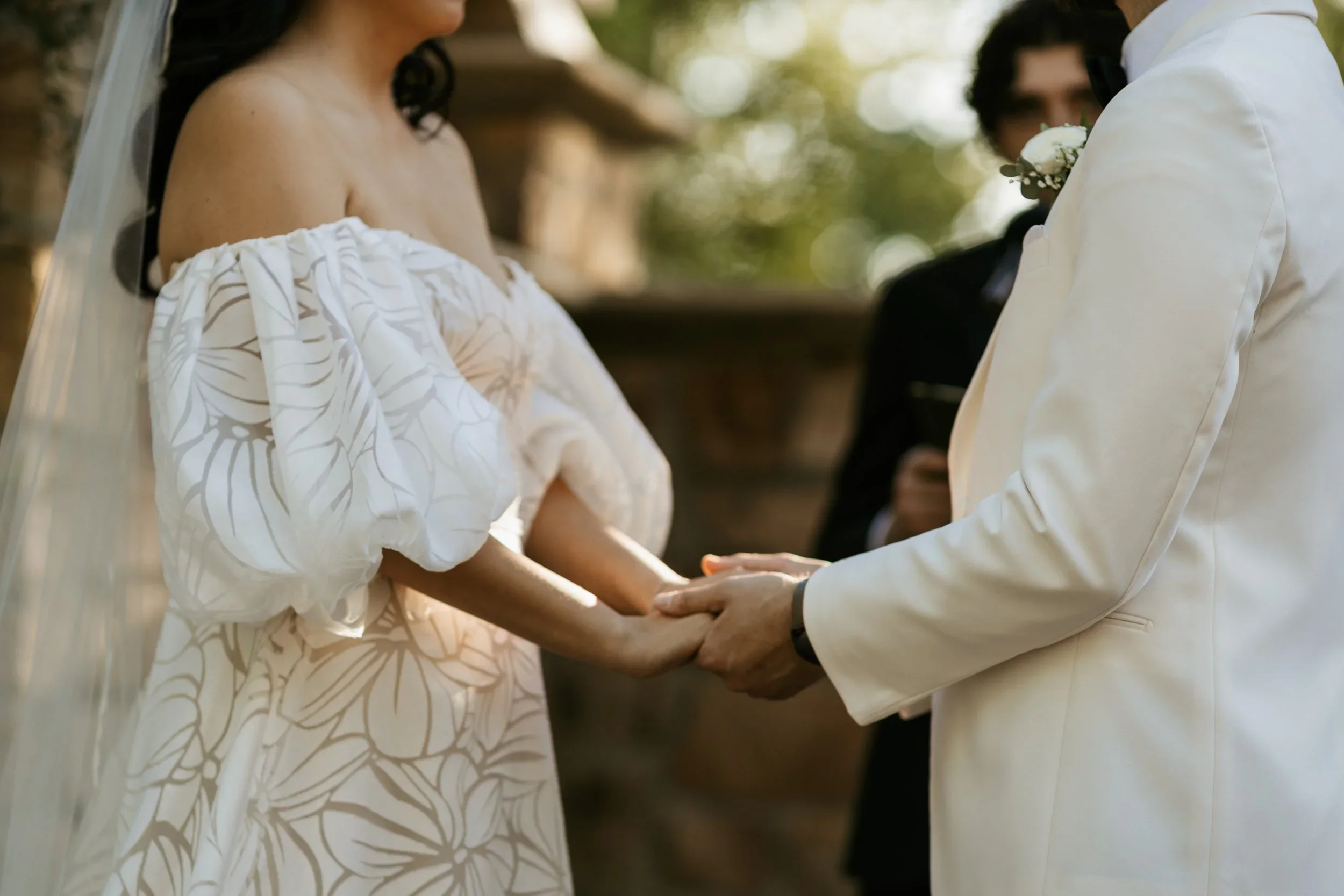 A bride and groom holding hands during their wedding ceremony, with an officiant in the background, outdoors under natural light.