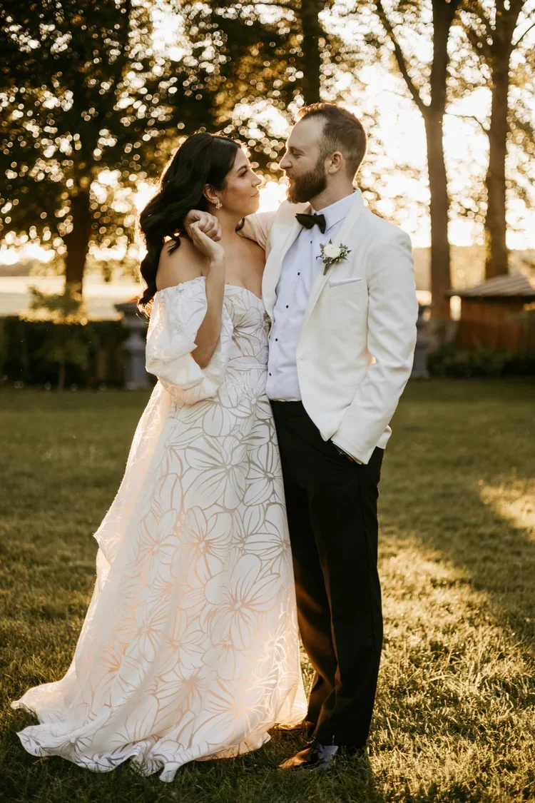 A bride and groom standing close together outdoors at sunset, looking into each other's eyes. The bride is wearing a white off-the-shoulder dress with a floral pattern, and the groom is wearing a white tuxedo jacket with black pants and a black bow t