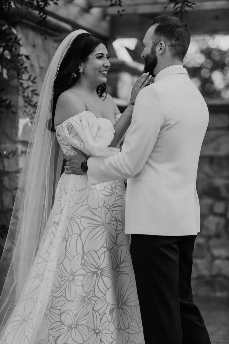 A black and white photo of a bride and groom smiling at each other during their wedding. The bride is wearing an off-the-shoulder dress with a leafy pattern, a veil, and earrings. The groom is wearing a white jacket with dark pants. The bride and gro