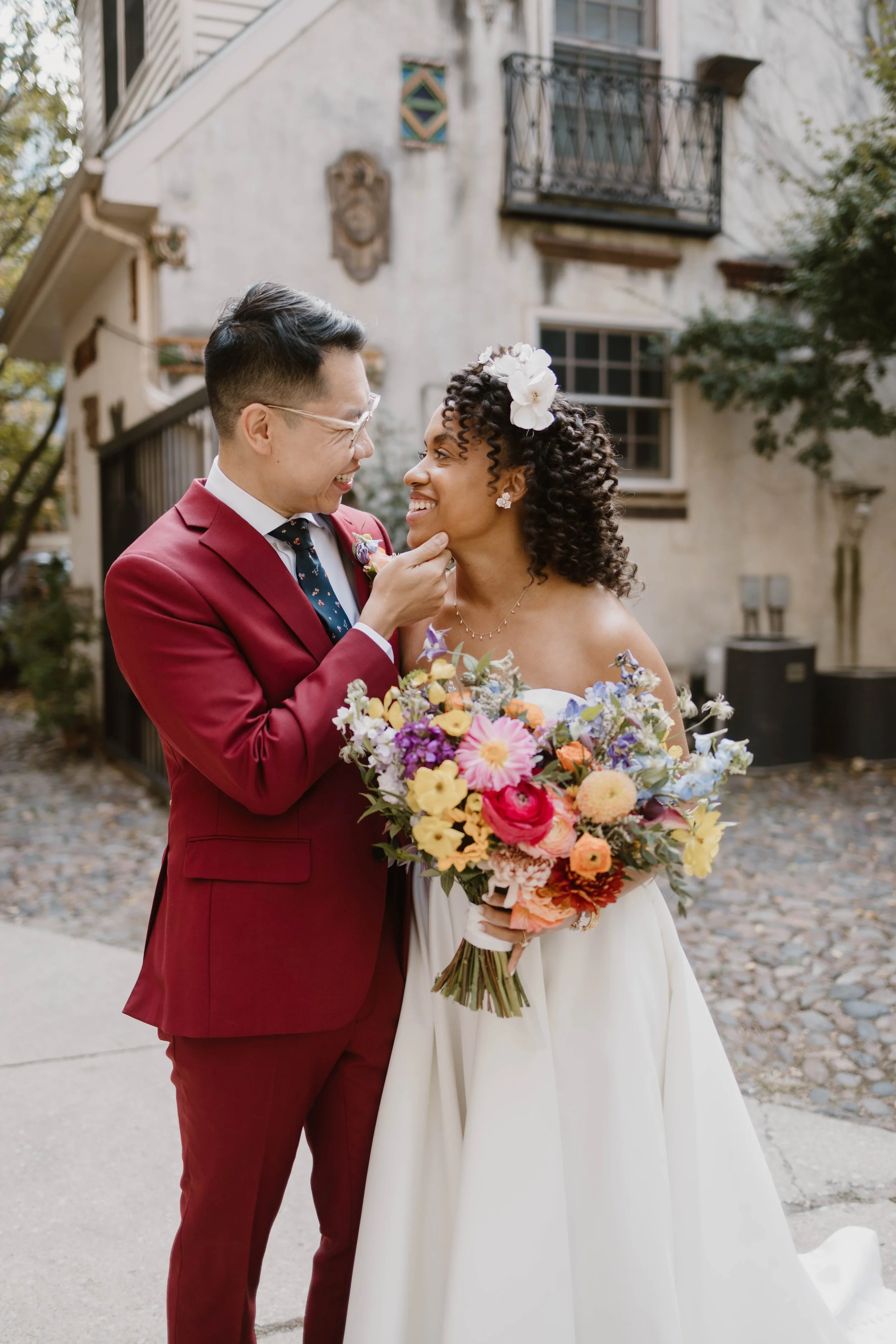 A couple in wedding attire sharing a joyful moment outside, with the man touching the woman's chin and both smiling at each other. The woman holds a colorful bouquet and wears a white dress with floral accessories in her curly hair, while the man wea