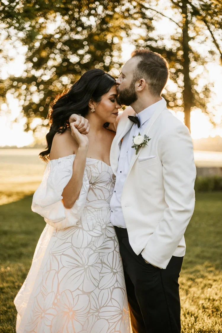 A bride and groom share a tender moment outdoors at sunset, with the groom gently kissing the bride's forehead while the bride has her eyes closed and a slight smile, both dressed in wedding attire.
