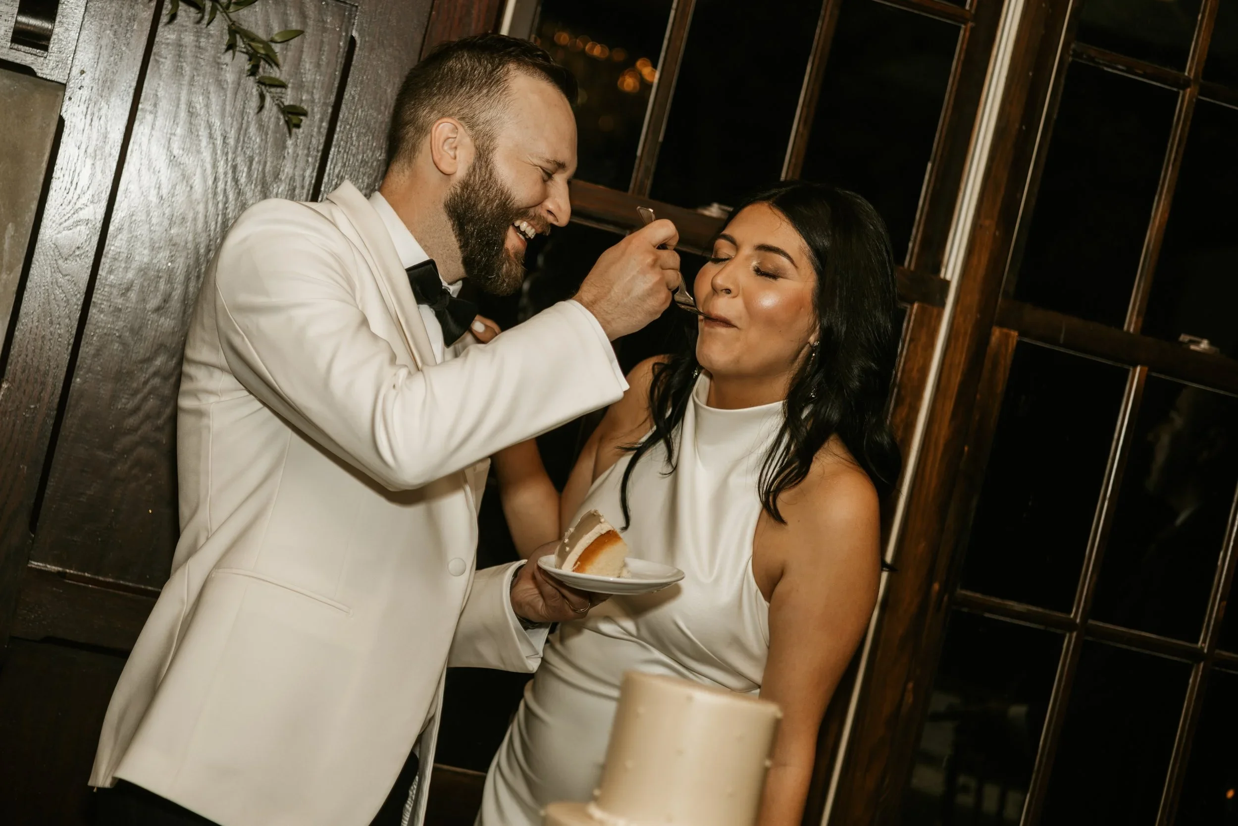 A man in a white tuxedo with a black bow tie feeds a woman in a white dress a piece of cake at a celebration or wedding.