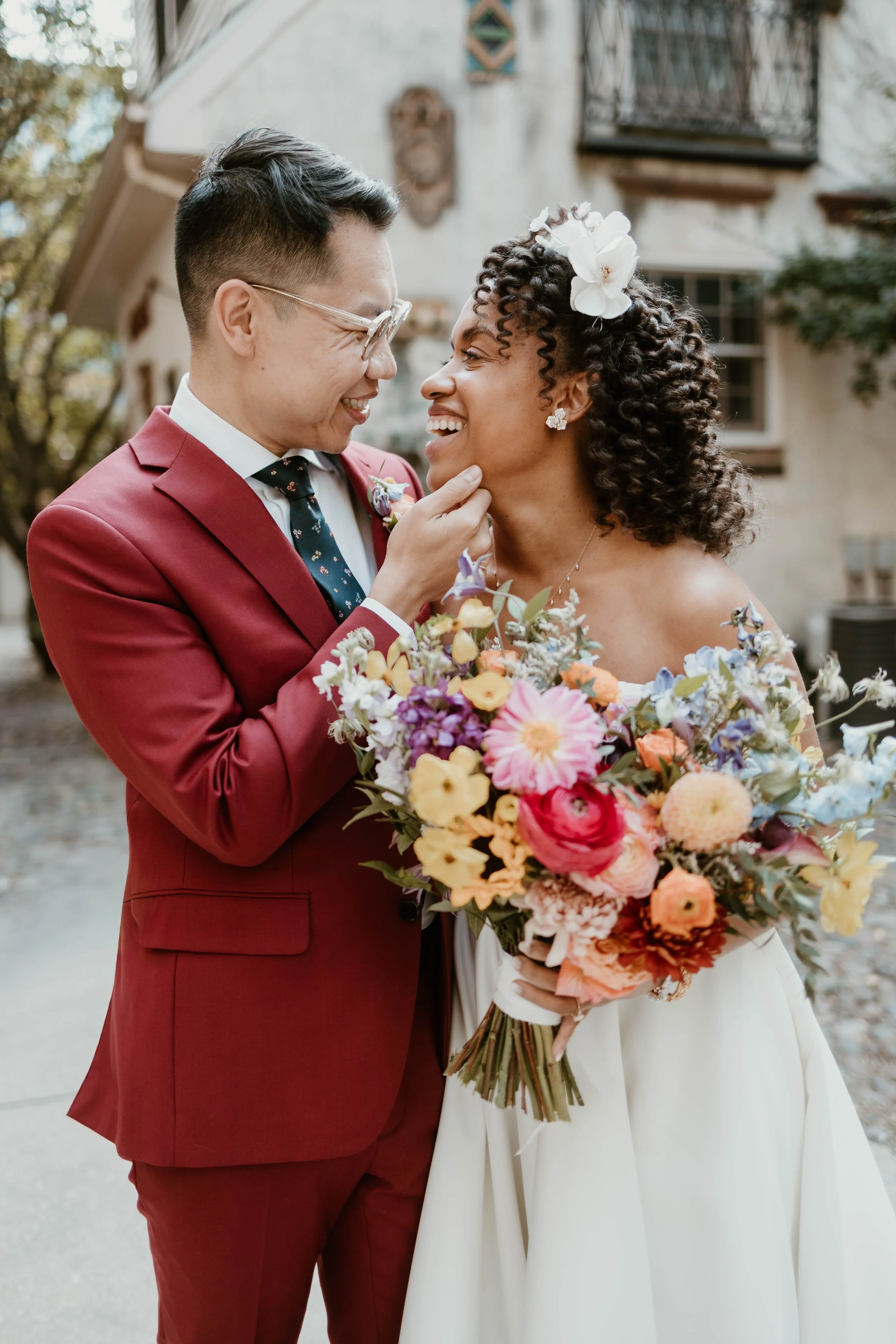 A wedding couple smiling and gazing at each other, the groom gently touching the bride's chin, with the bride holding a colorful bouquet of flowers, outdoors in front of a rustic building.