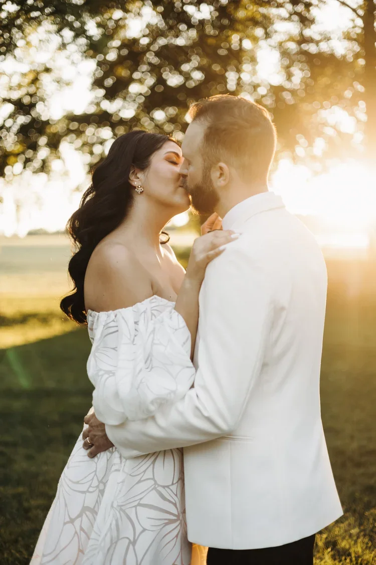 A couple in wedding attire sharing a kiss outdoors during sunset, with trees and sunlight in the background.