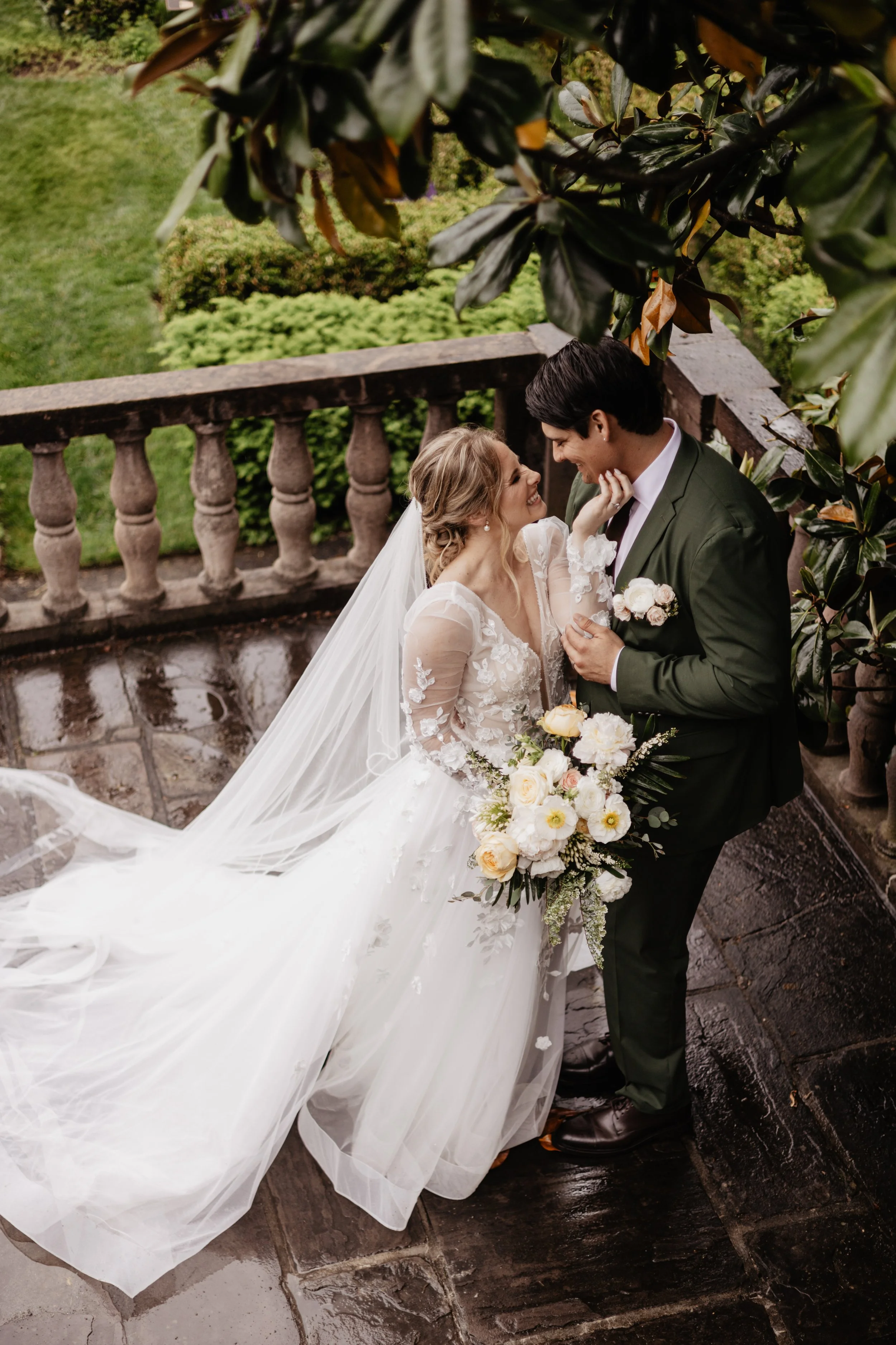 A bride and groom sharing a romantic moment outdoors on their wedding day, surrounded by greenery and stone railings.