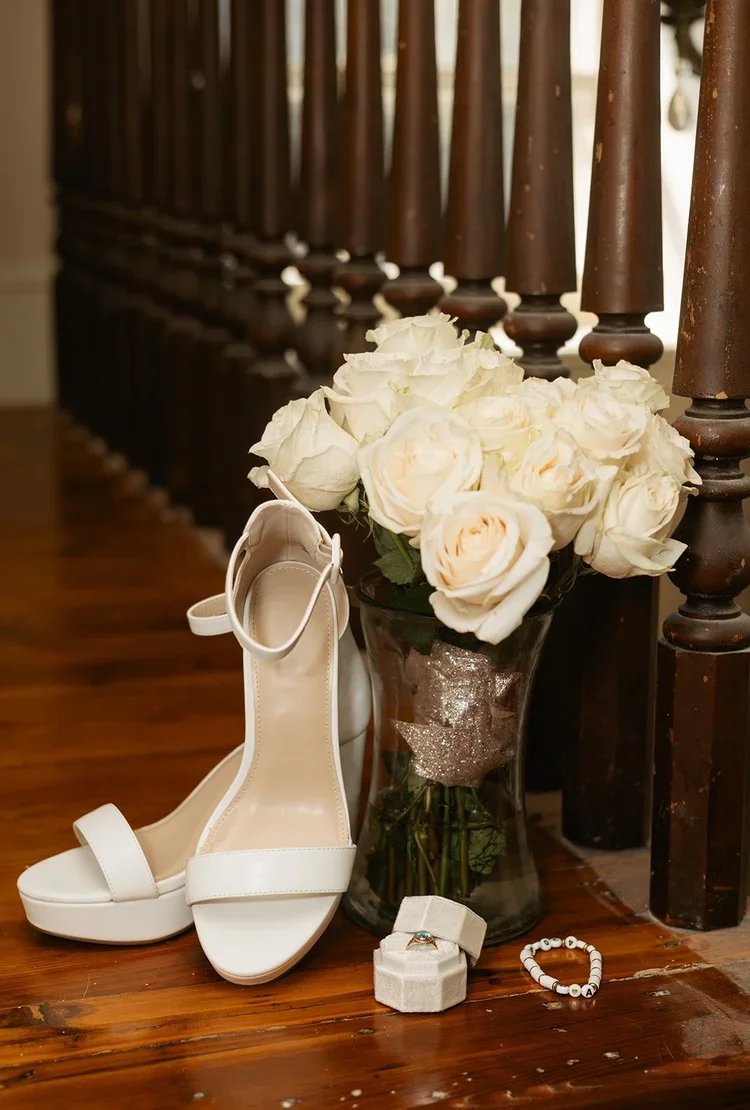 A pair of white high heel sandals, a bouquet of white roses in a glass vase, a ring in a small box, and a beaded bracelet placed on a wooden floor next to a staircase.