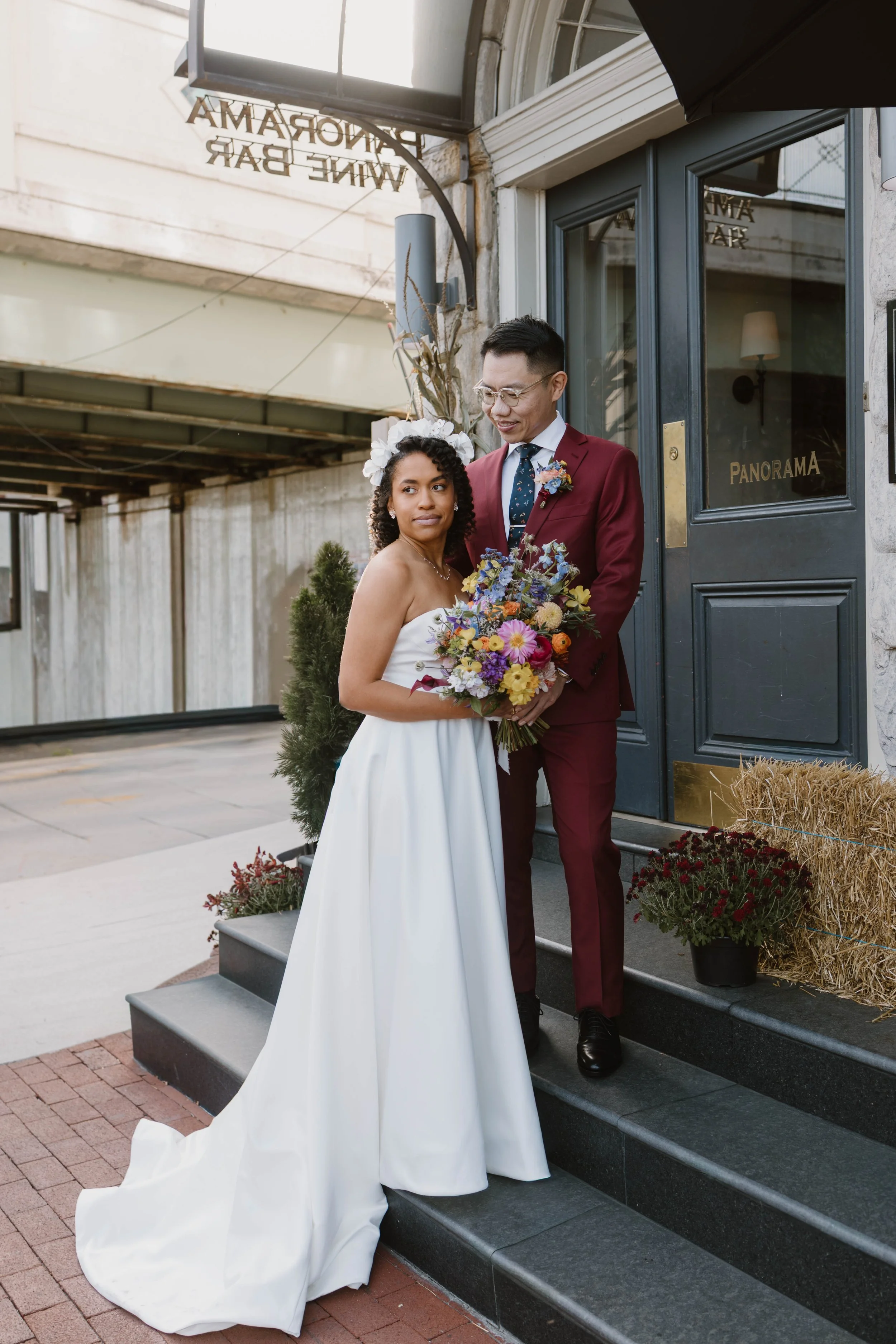 A bride and groom standing on the steps outside a building labeled 'Panorama.' The bride is in a white wedding dress holding a colorful bouquet, and the groom is in a burgundy suit with a floral tie and boutonniere, smiling at each other.