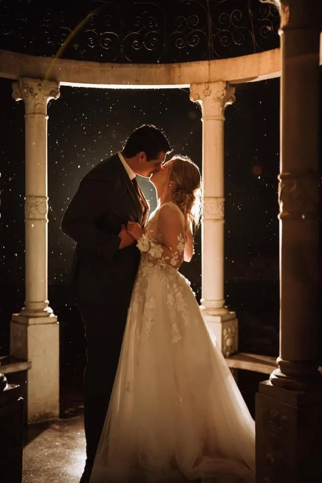 A bride and groom sharing a kiss under a gazebo at night, with rain falling around them.