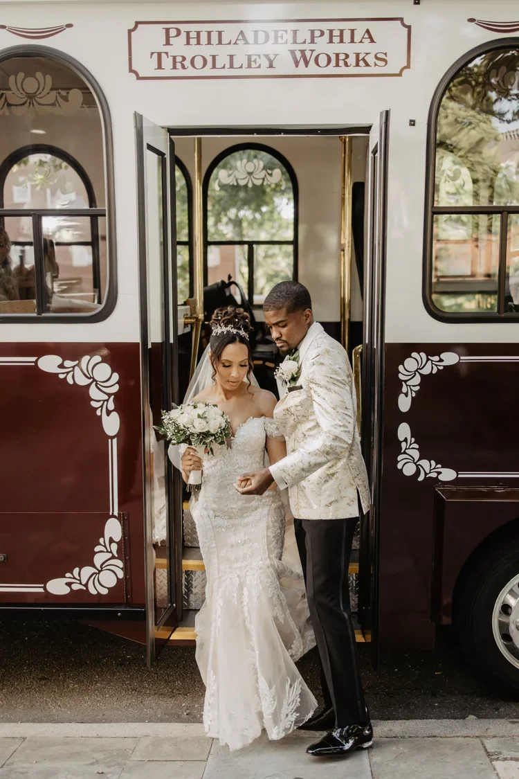 A bride and groom standing outside a trolleybus labeled 'Philadelphia Trolley Works', with the bride holding a bouquet of white flowers, and the groom adjusting a bracelet on her wrist