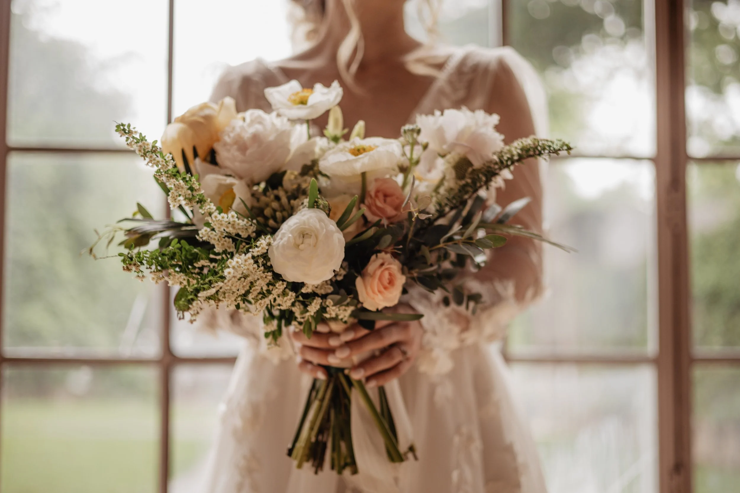 A bride in a wedding dress holding a bouquet of white, pink, and cream flowers, standing near large windows with nature visible outside.