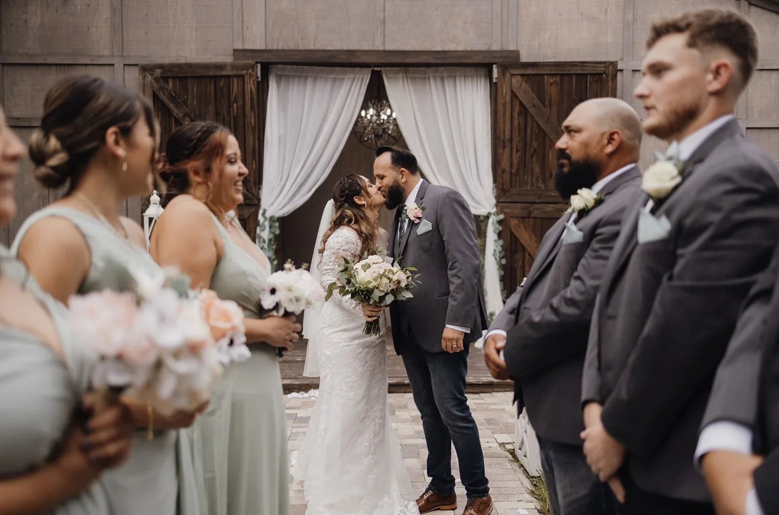Couple sharing a kiss during their wedding ceremony, surrounded by bridesmaids and groomsmen, with rustic barn background.