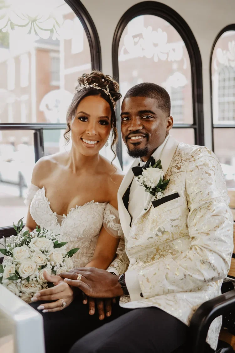 A bride and groom sitting together at their wedding, smiling and holding hands indoors near large windows.