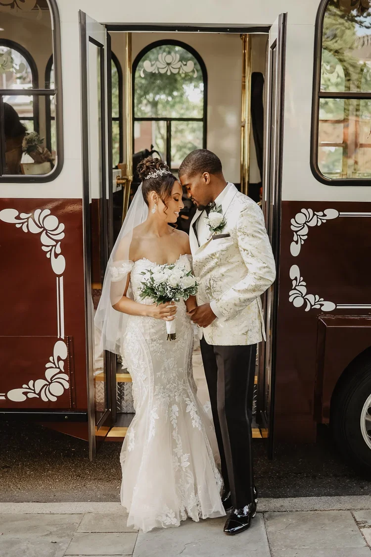 A bride and groom sharing a moment outside a vintage bus, standing close with foreheads touching, dressed in wedding attire. The bride holds a bouquet and wears a lace wedding gown with an off-the-shoulder design, while the groom wears a white tuxedo