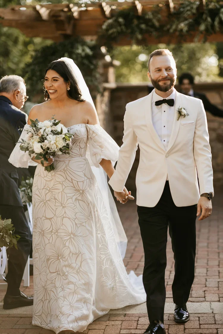 A newlywed couple holding hands at their outdoor wedding, with the bride in a white floral dress holding a bouquet and the groom in a white tuxedo jacket, black bow tie, and black pants, smiling as they walk.