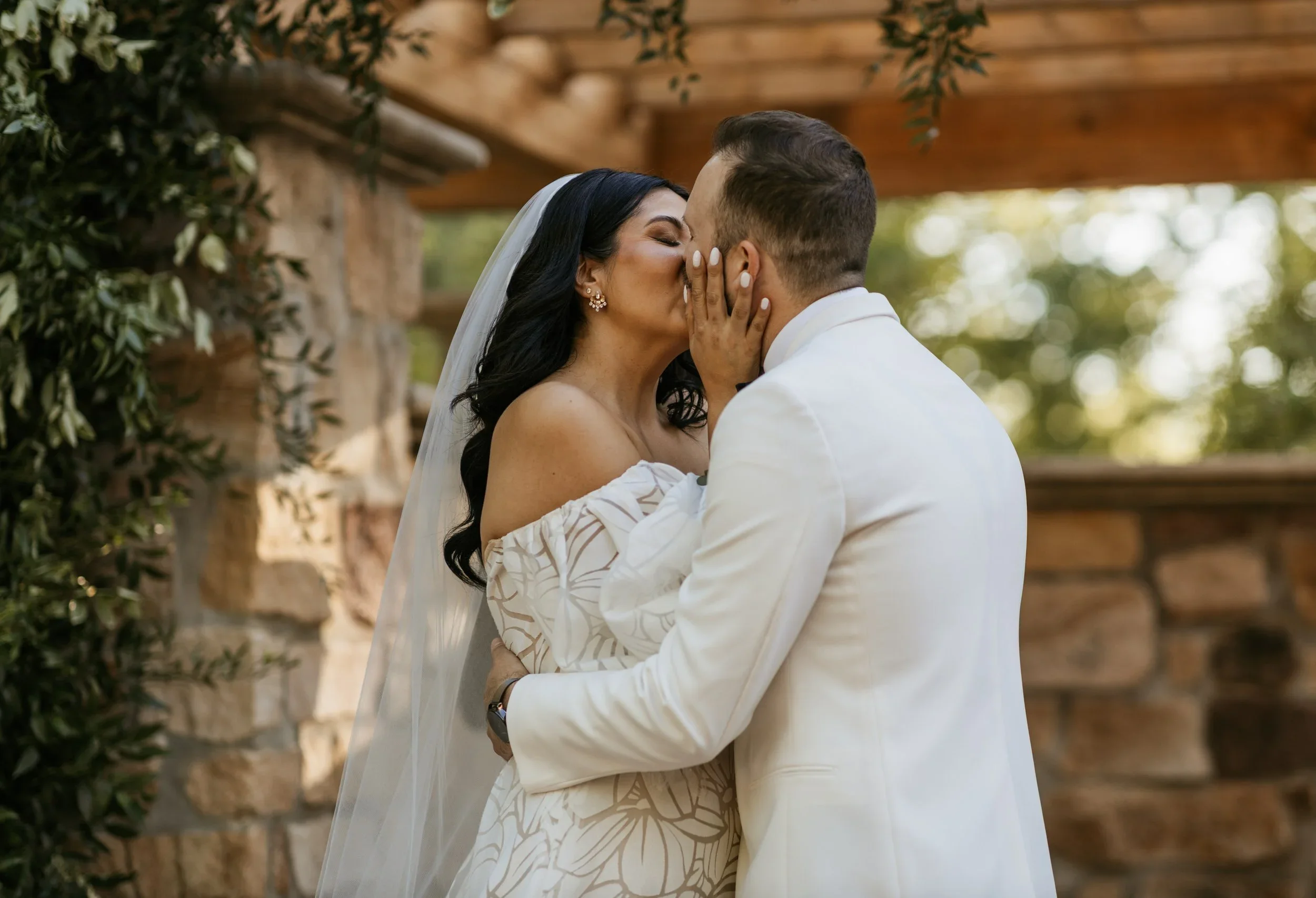 A couple sharing a kiss at their wedding outdoors, with greenery and a brick wall in the background.