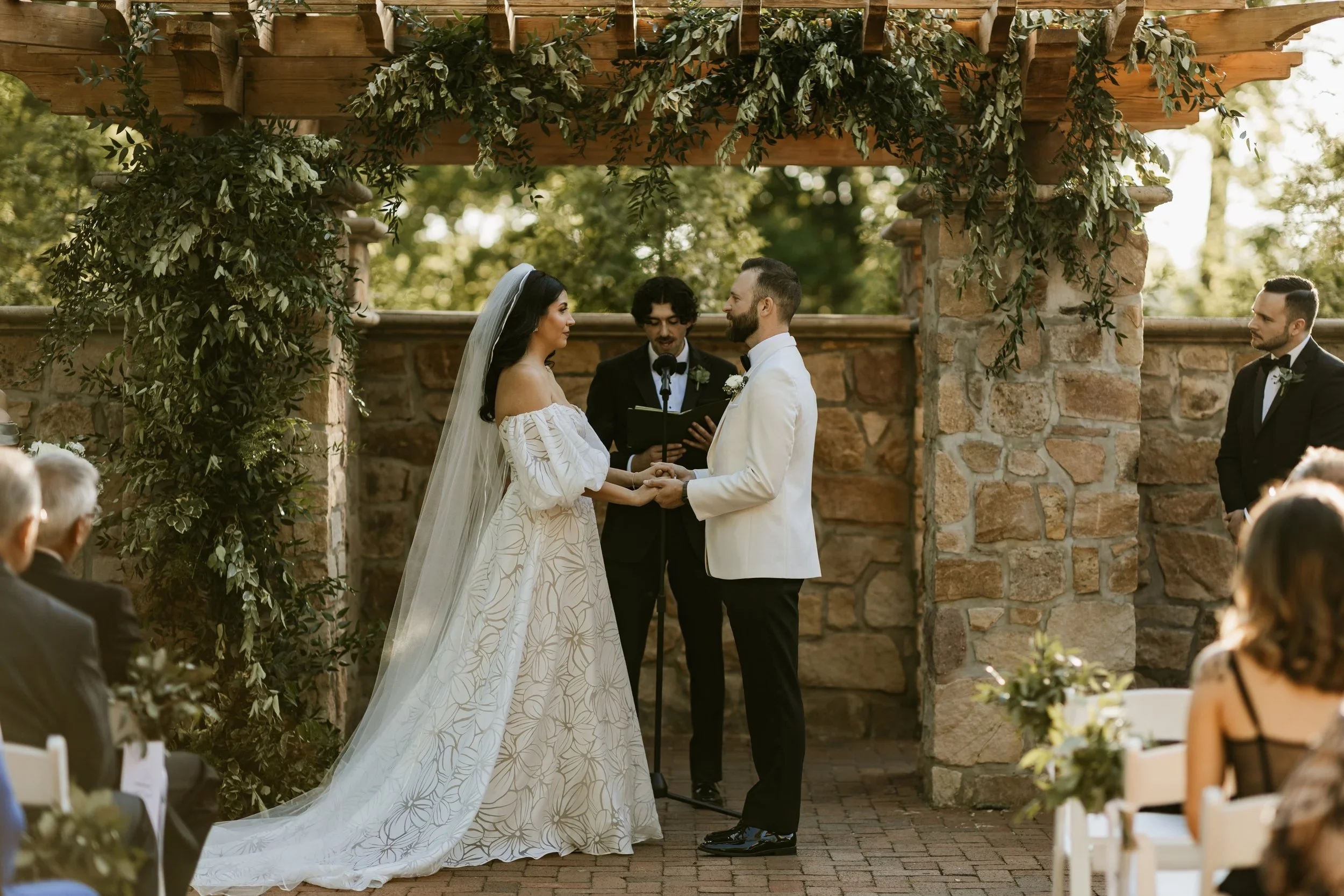A couple getting married outdoors under a wooden arch decorated with greenery, holding hands and exchanging vows. The bride is wearing a white off-shoulder wedding gown and veil, and the groom is dressed in a white tuxedo with black pants. An officia