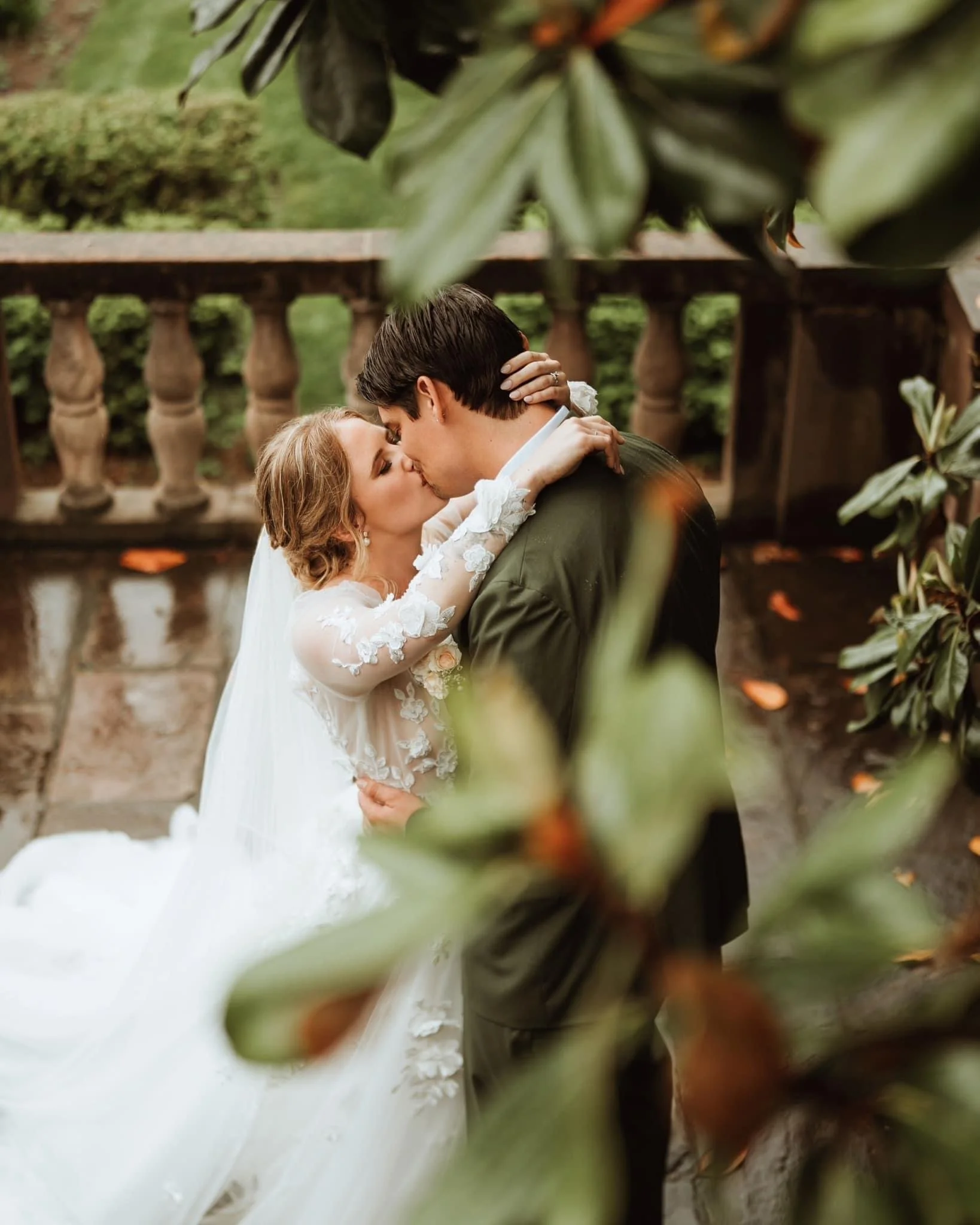 A bride and groom sharing a kiss outdoors, with greenery and a stone railing in the background, framed by leaves in the foreground.