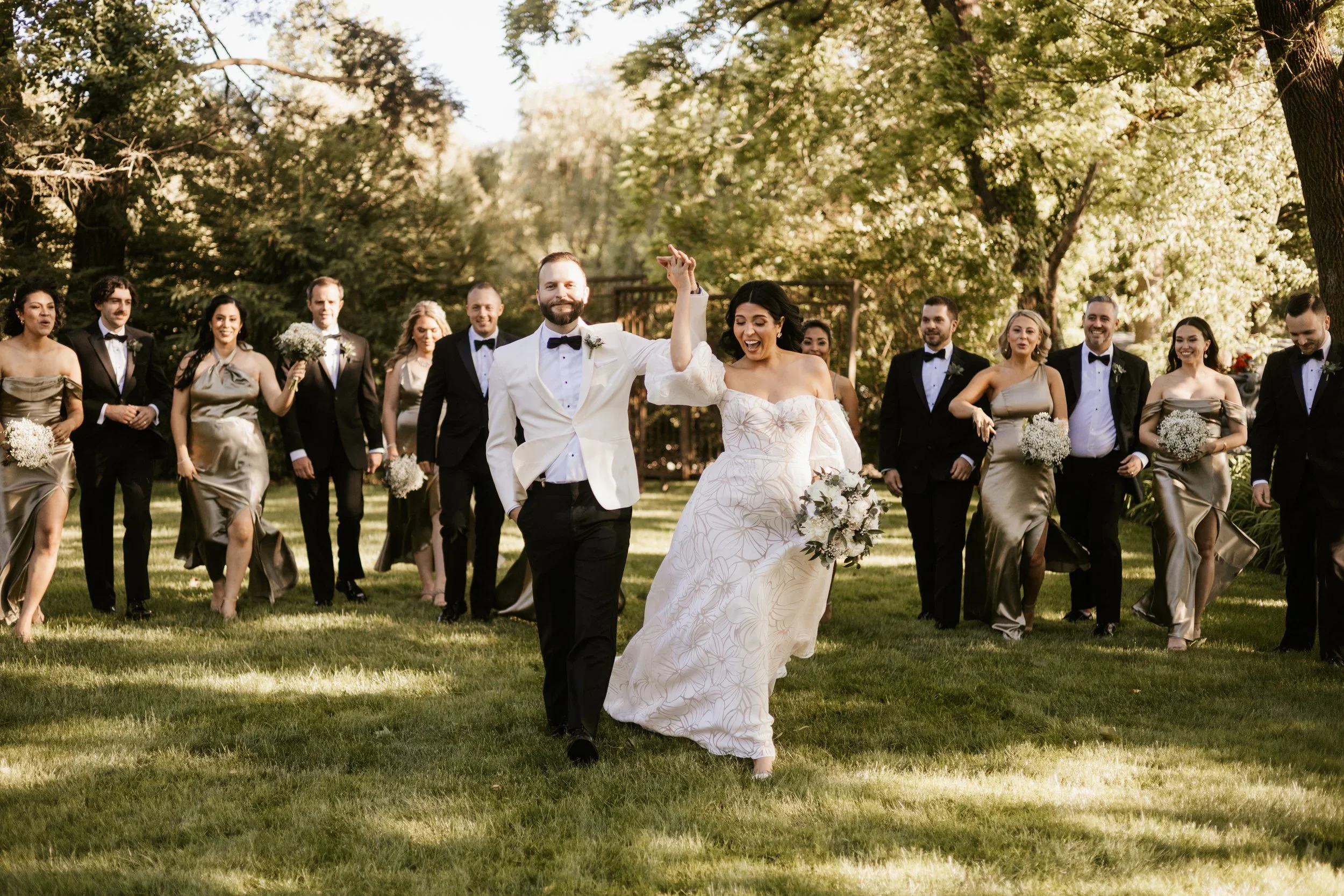 A wedding couple, the groom in a white tuxedo jacket and the bride in a white dress, walking hand-in-hand with friends and family in formal attire in a park with trees and sunlight.