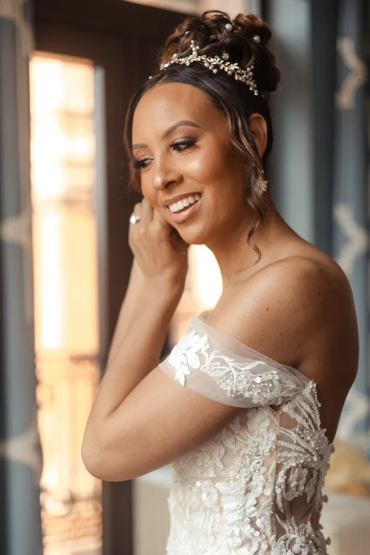 A bride with dark hair styled in an updo wearing a pearl and floral headpiece. She is smiling and adjusting her earring, dressed in an elegant off-shoulder lace wedding gown with intricate embroidery.