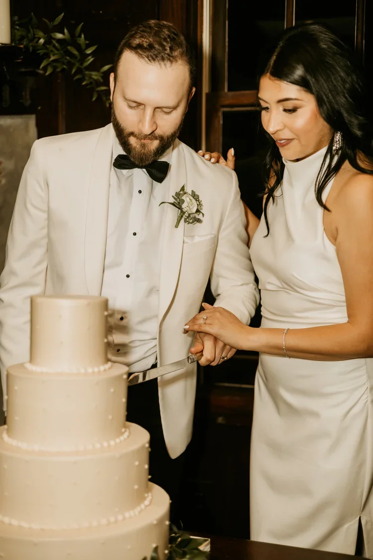 A newlywed couple dressed in a tuxedo and a white dress cutting a wedding cake together at their celebration.