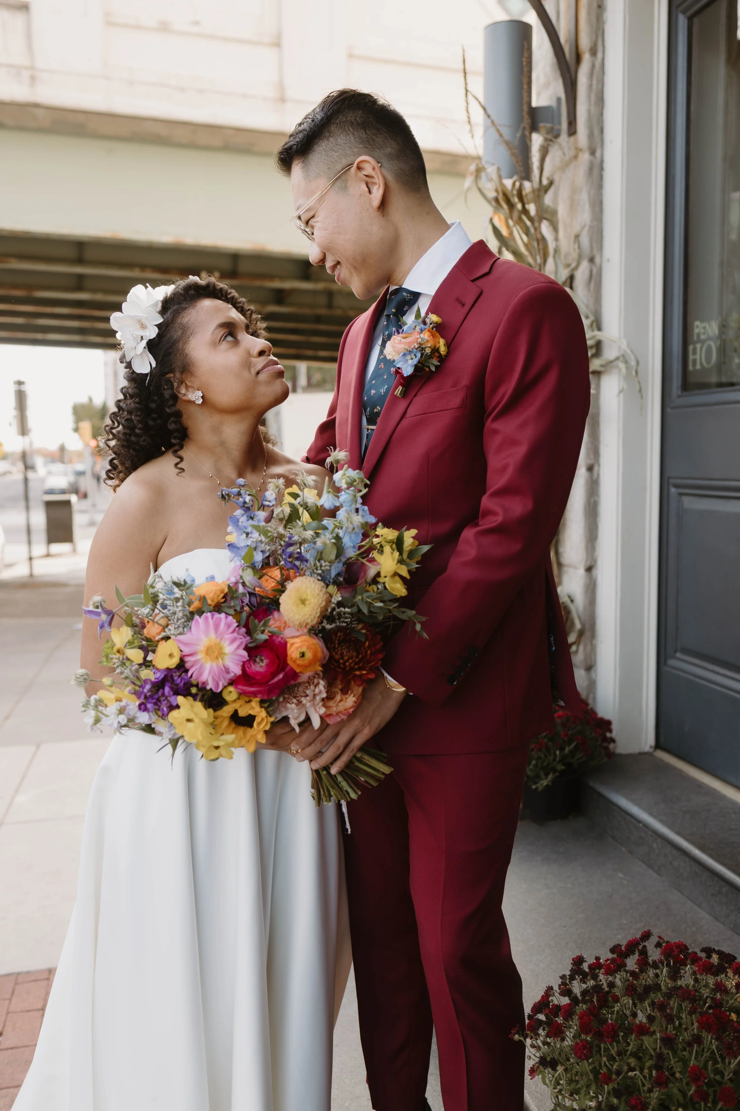 A bride and groom stand close together outdoors, gazing at each other. The bride holds a colorful bouquet of flowers and is dressed in a strapless white gown with a floral headpiece. The groom wears a burgundy suit with a floral boutonniere and glass