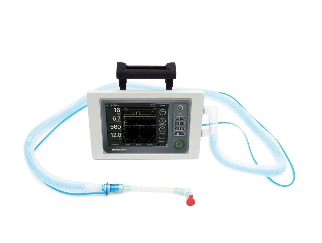 Photograph of iSimulate's portable Hamilton T1 medical ventilator simulator with display showing respiratory data and controls, and various hoses and attachments extending from the right-hand panel.