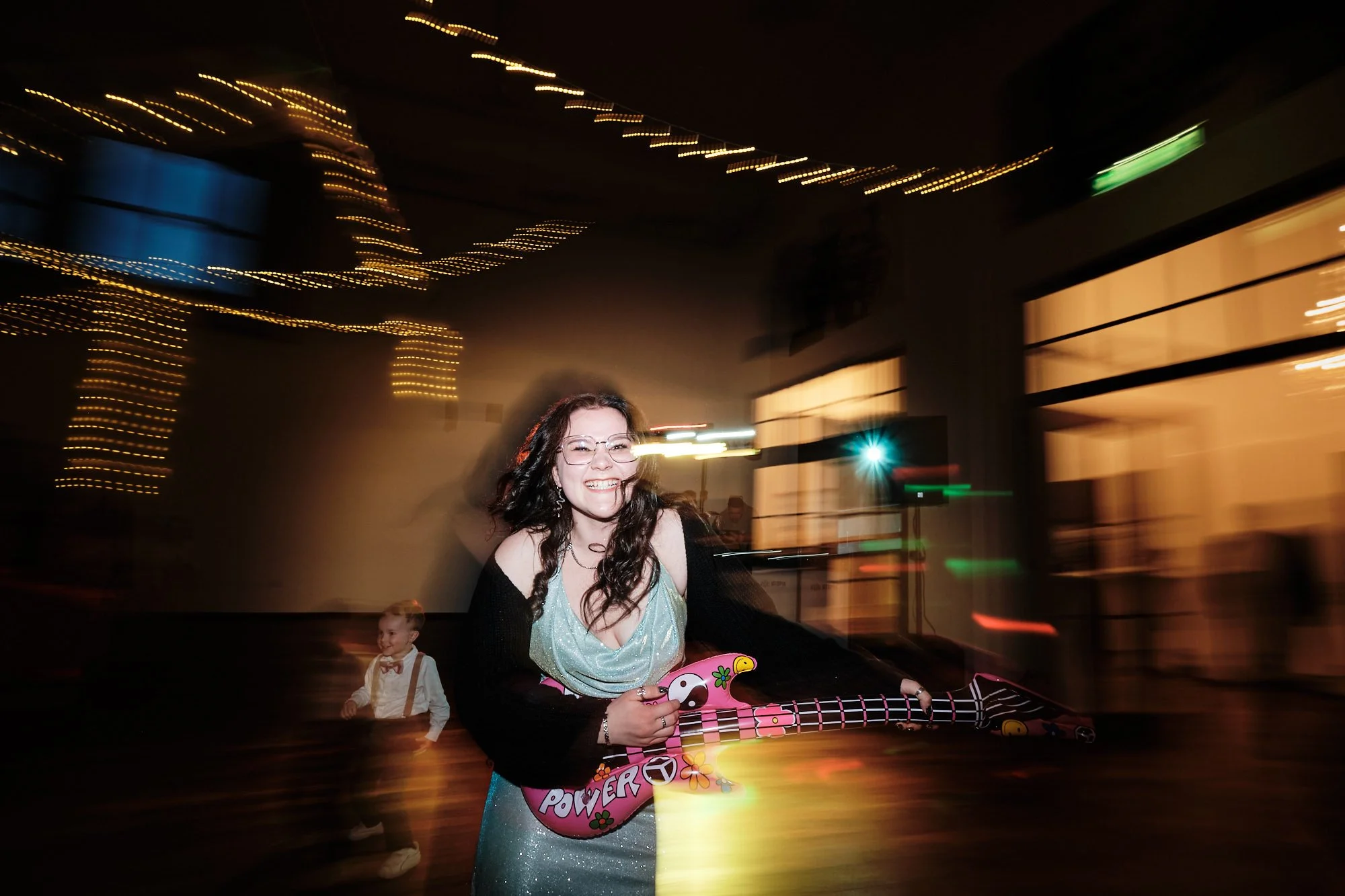 Hochzeitsparty: fröhliche Frau in glitzerndem Kleid mit buntem Spielzeug-Gitarre, tanzt vor Lichterketten; Kind im Hintergrund.