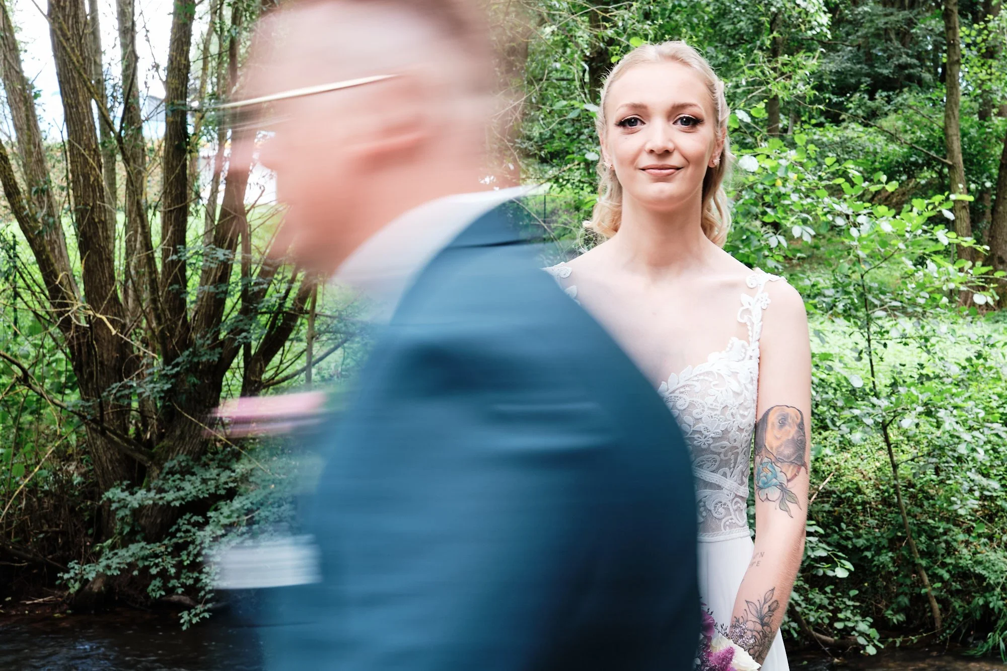 künstlerische Aufnahme: Blick der Braut in die Kamera, während der Bräutigam mit Bewegungsunschärfe vorbeizieht; Waldkulisse.