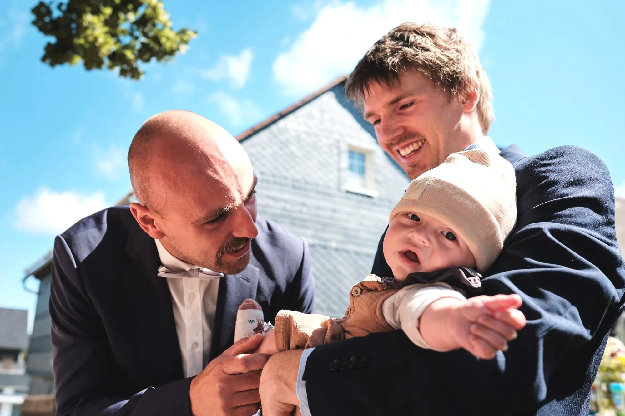 Zwei Männer halten einen Baby in der Hand, alle lachen im Freien bei blauem Himmel.