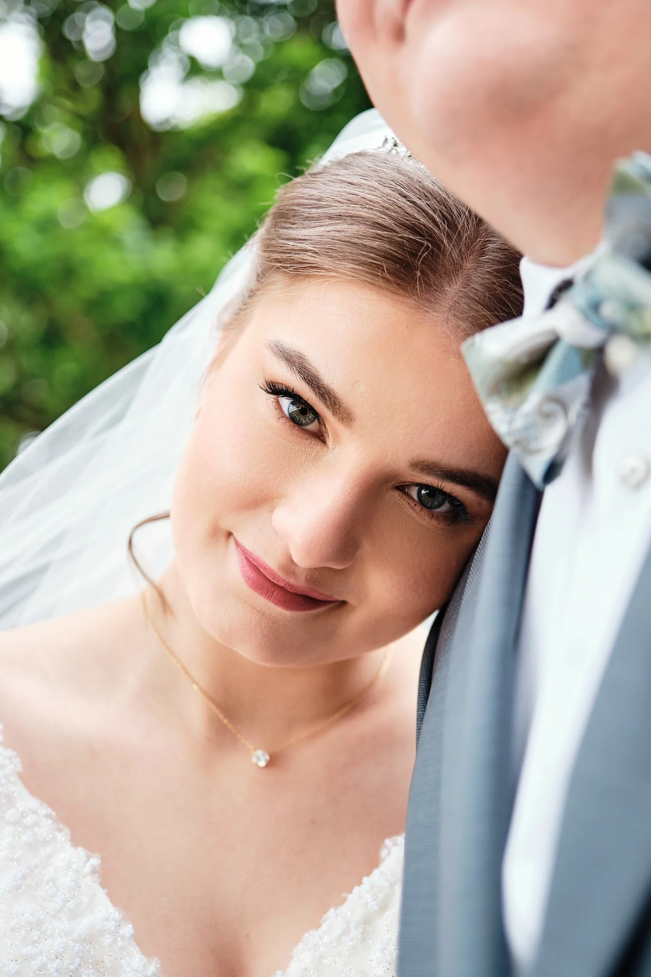 Nahaufnahme eines Brautpaares bei Hochzeit, die Braut lächelt und lehnt ihren Kopf an die Schulter des Bräutigams, im Hintergrund grüne Blätter.