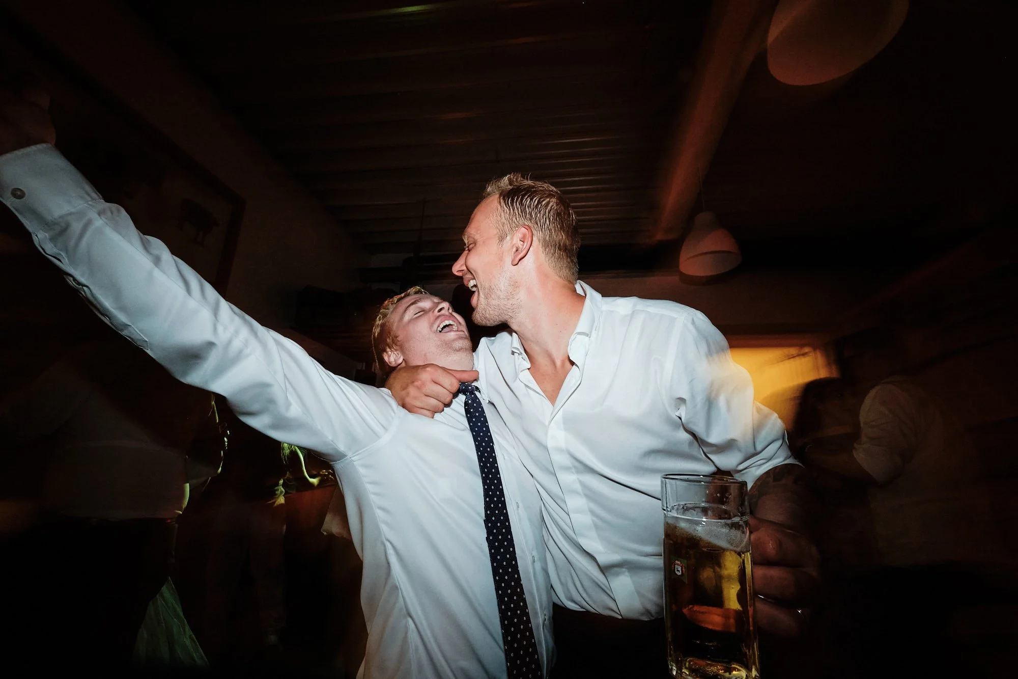 Hochzeitsparty: Bräutigam und Gast feiern ausgelassen, Arm in Arm, Bierglas in der Hand.